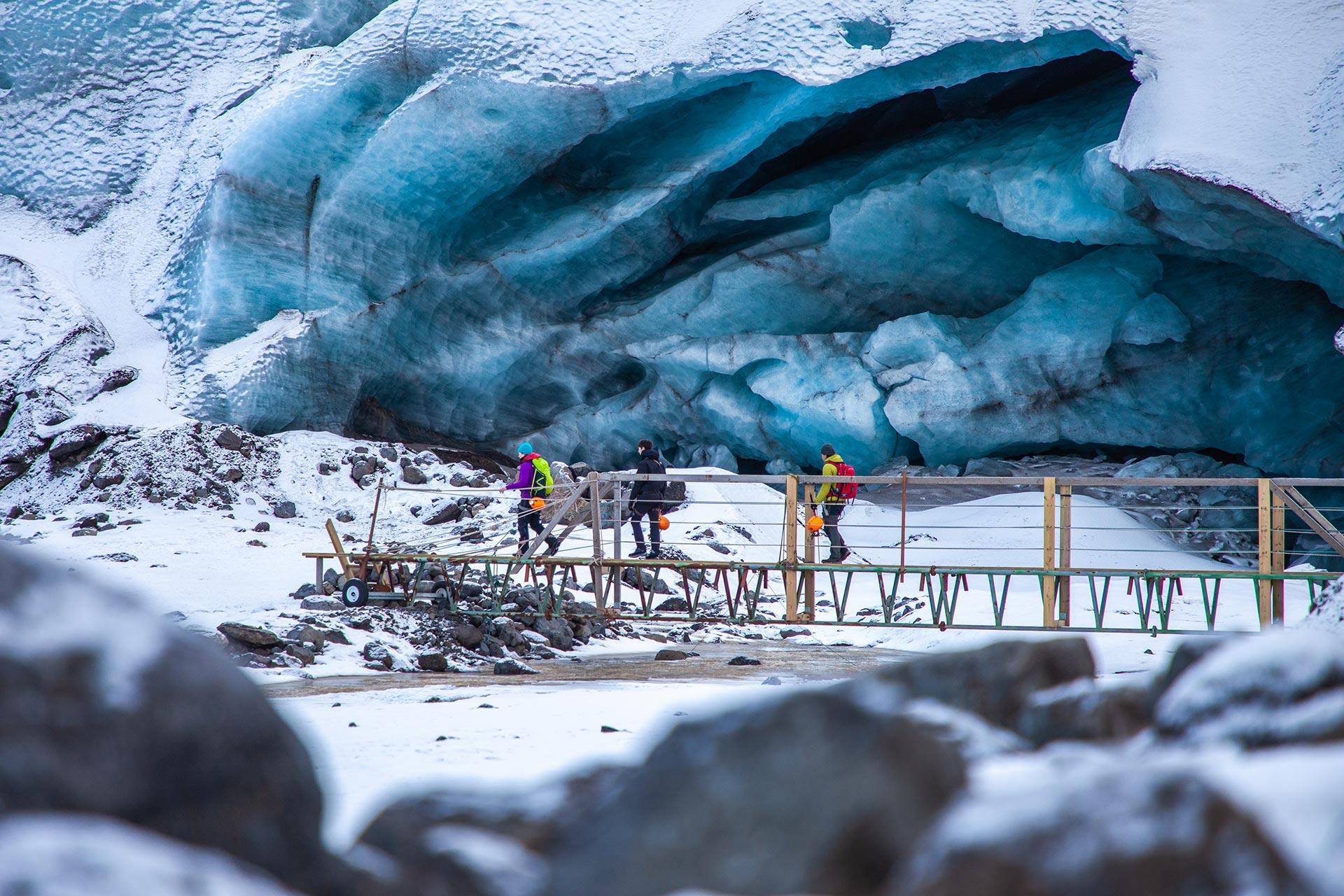 People walking over a bridge past an ice cave that is very blue