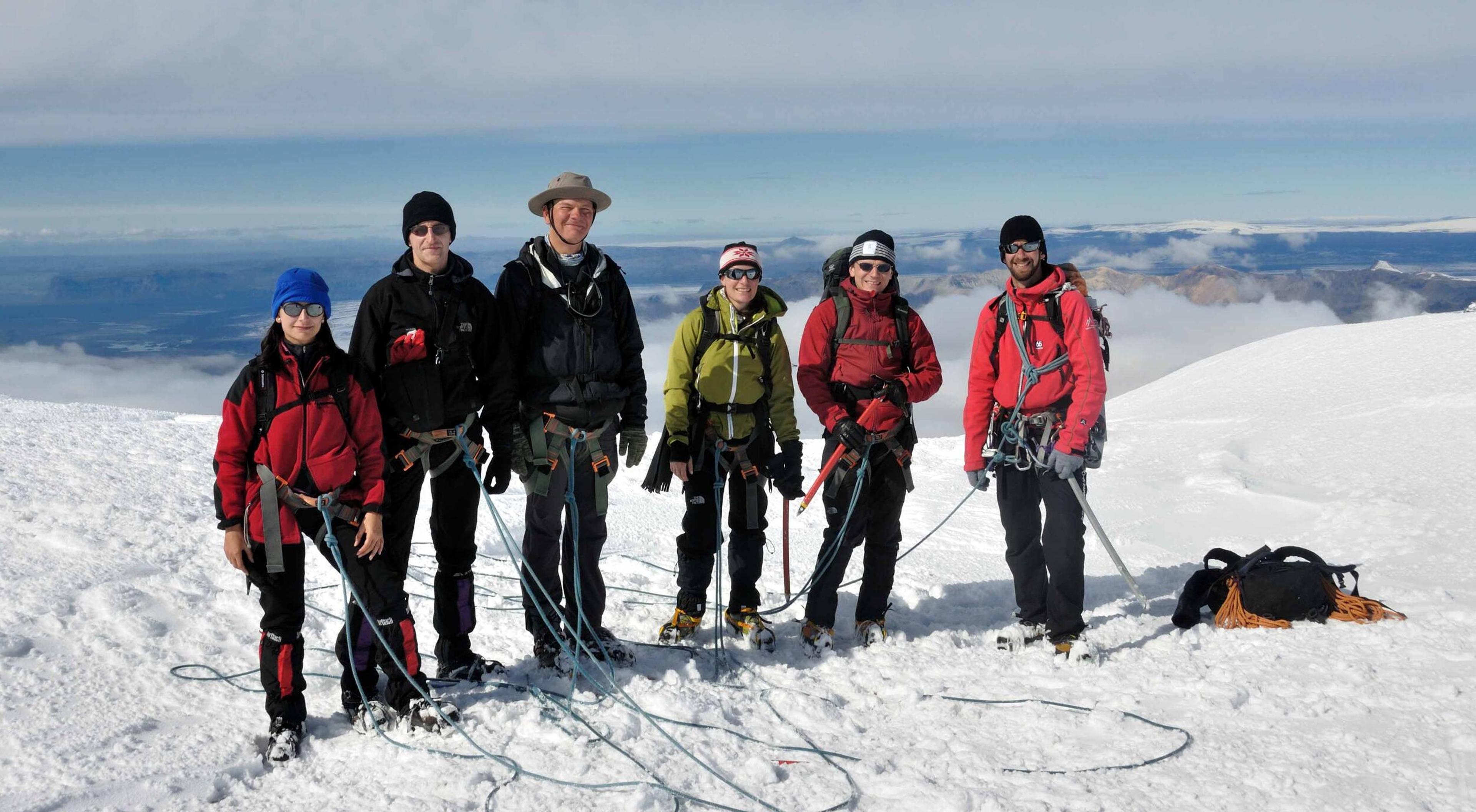 A group of six hikers, equipped with climbing gear, stands together on the snowy summit of Eyjafjallajökull glacier under a bright, clear sky.