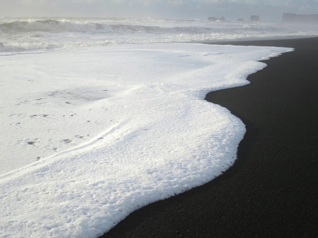 Foaming waves and the black sand contacting with the Dyrholaey stone arc in the background