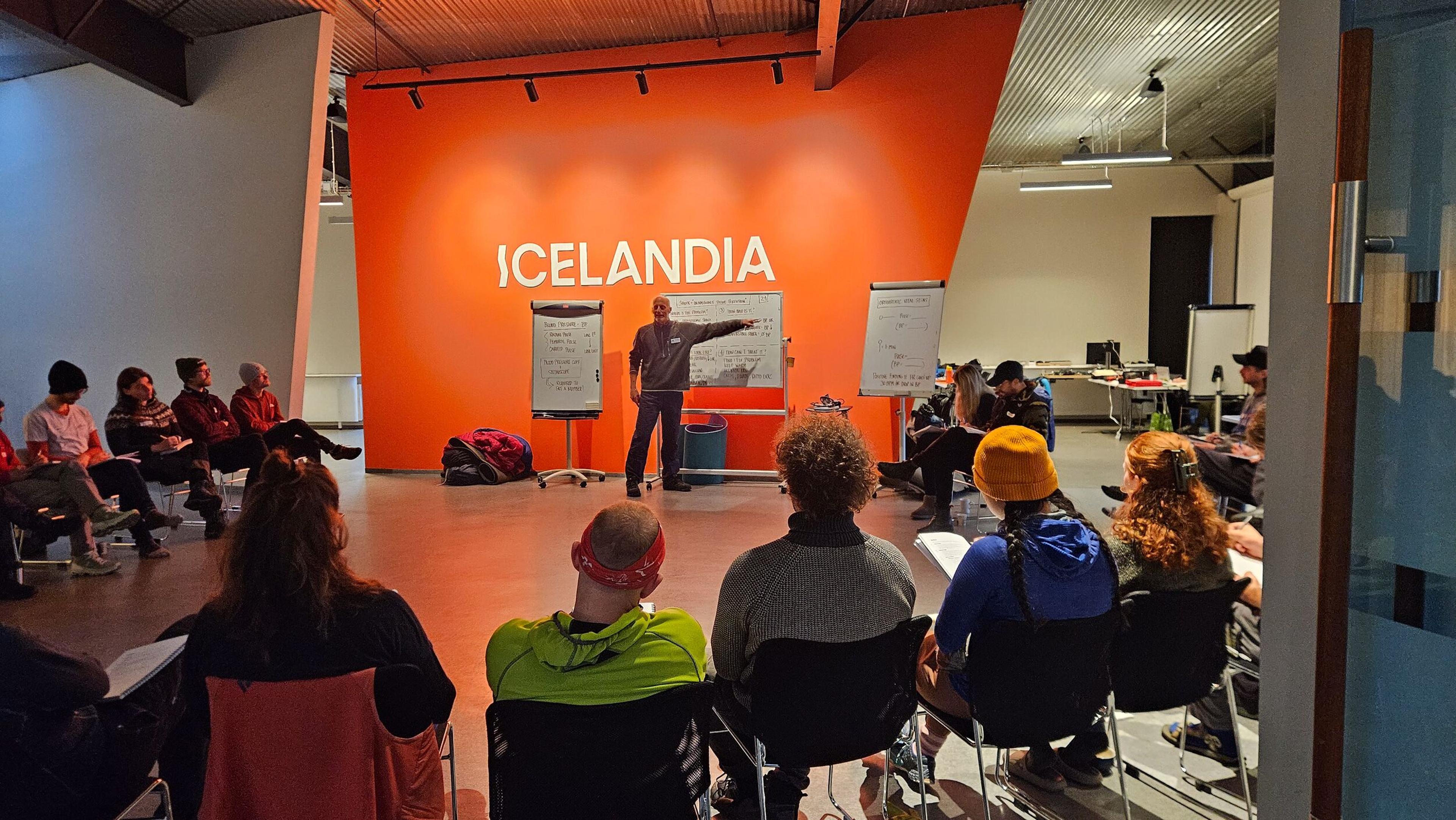  a workshop or training session held in a room with a bright orange wall labeled "ICELANDIA." Participants are seated in a semi-circle, listening to a presenter who stands in front of a whiteboard and presentation materials