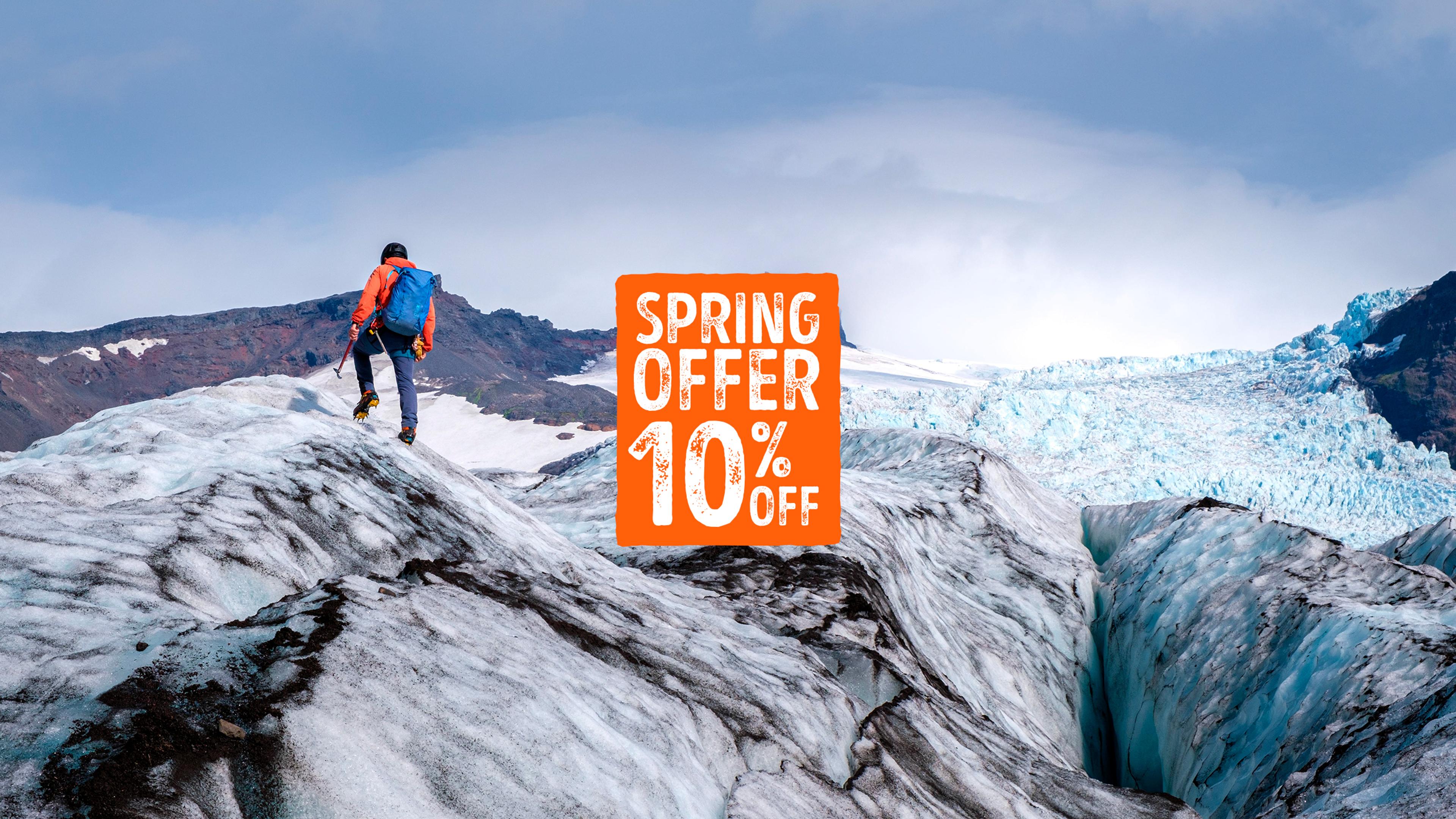 This image shows a person wearing colorful pants, a backpack, and a bright orange beanie hiking across a stunning glacier. The icy blue surface of the glacier contrasts with the dark mountains and snow-capped peaks in the background, evoking a sense of adventure in a remote and frigid landscape