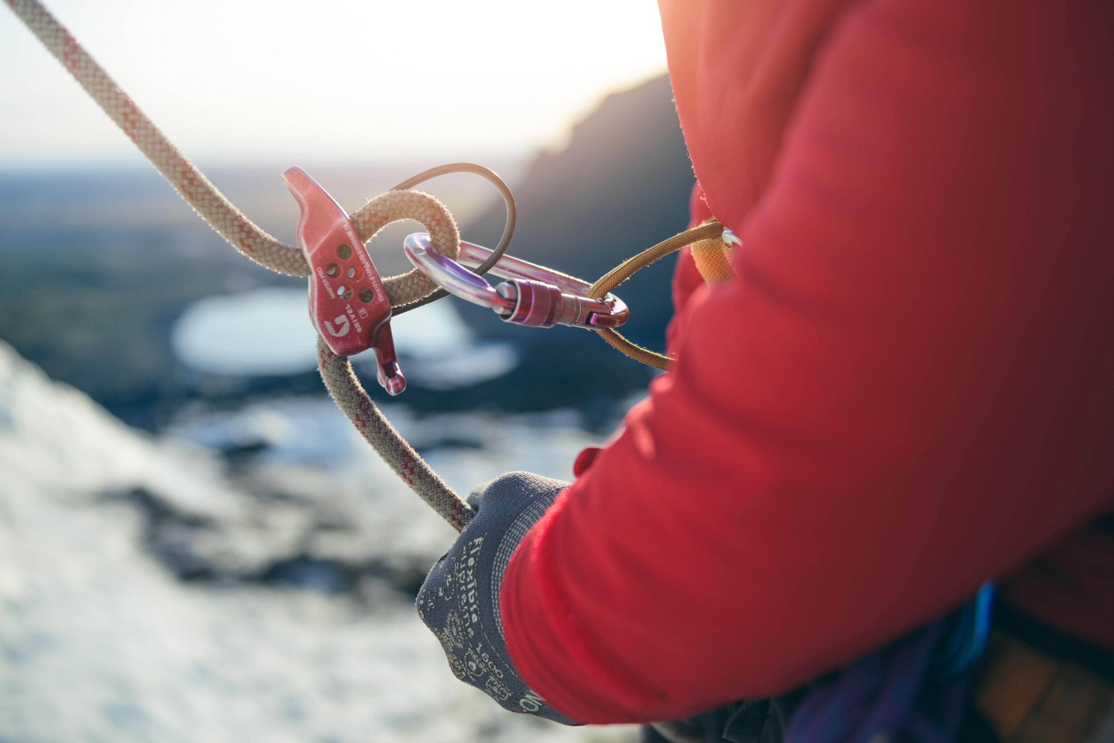"Close-up of a hand in a climbing glove holding a rope attached to a belay device and carabiner, with a glacial landscape blurred in the background