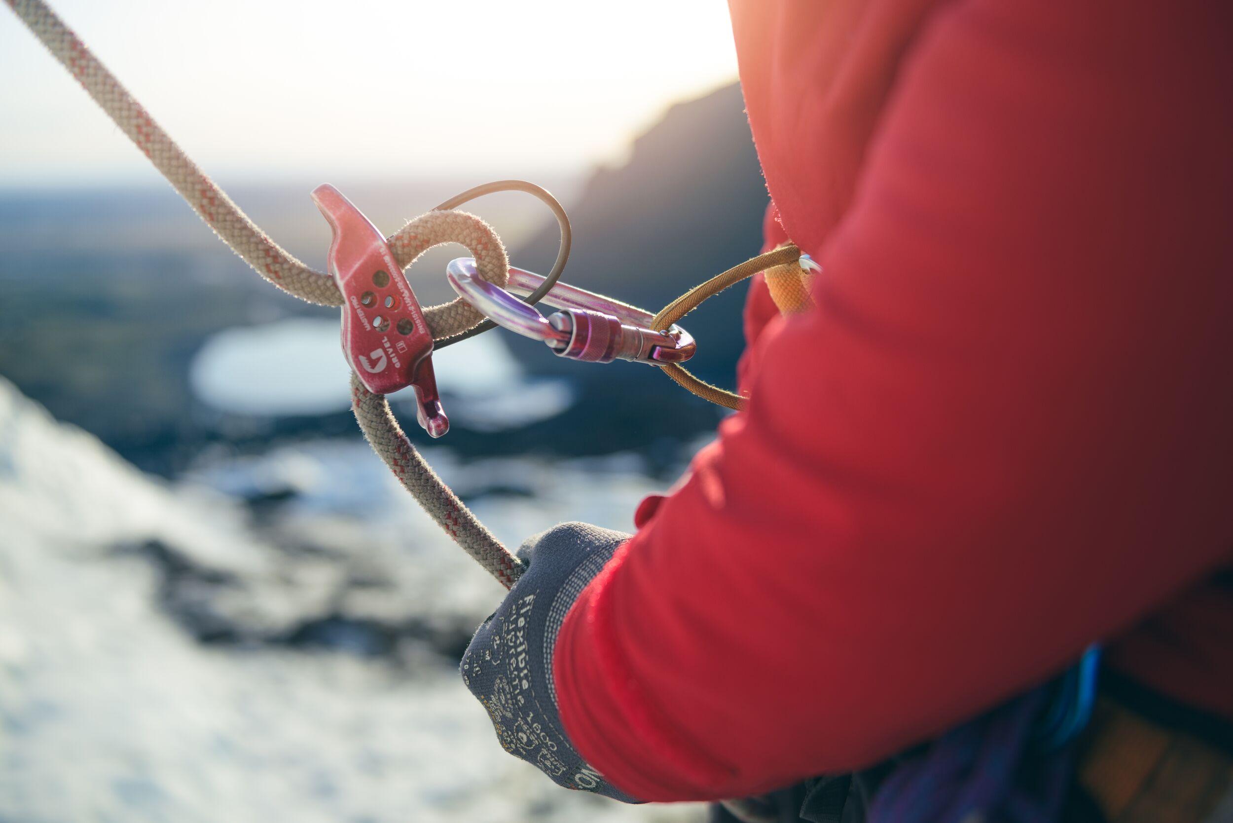 "Close-up of a hand in a climbing glove holding a rope attached to a belay device and carabiner, with a glacial landscape blurred in the background