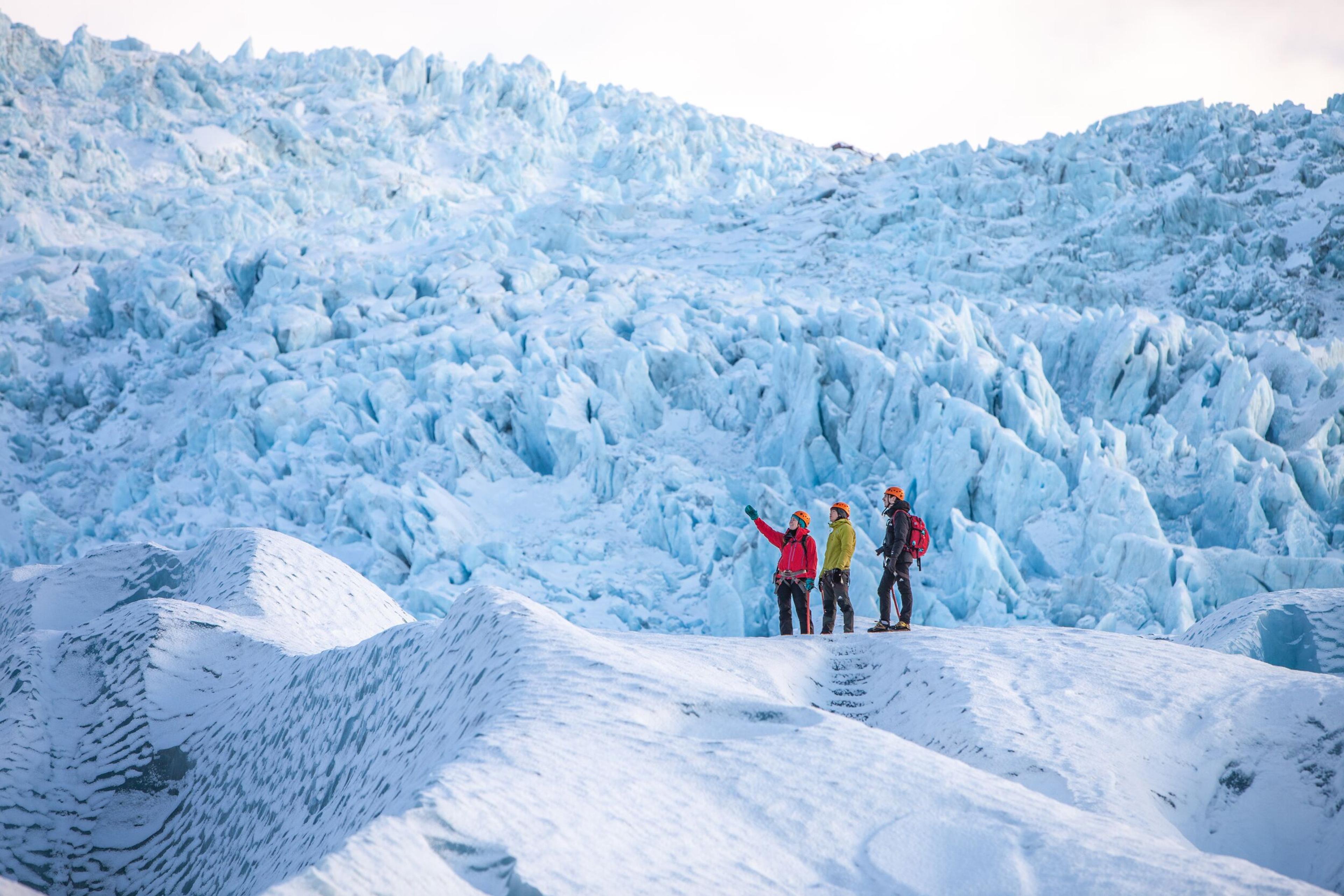 Three hikers stand atop a glacier ridge, one pointing upward, dwarfed by a massive icefall of jagged blue-white ice rising before them