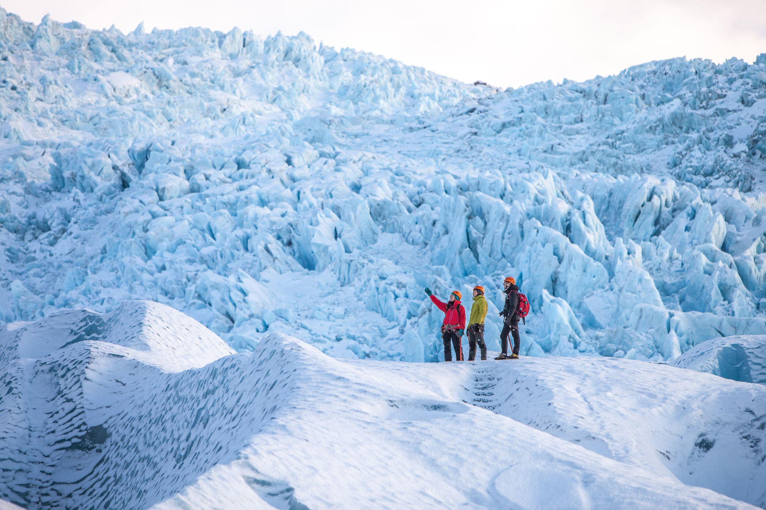 Three hikers stand atop a glacier ridge, one pointing upward, dwarfed by a massive icefall of jagged blue-white ice rising before them