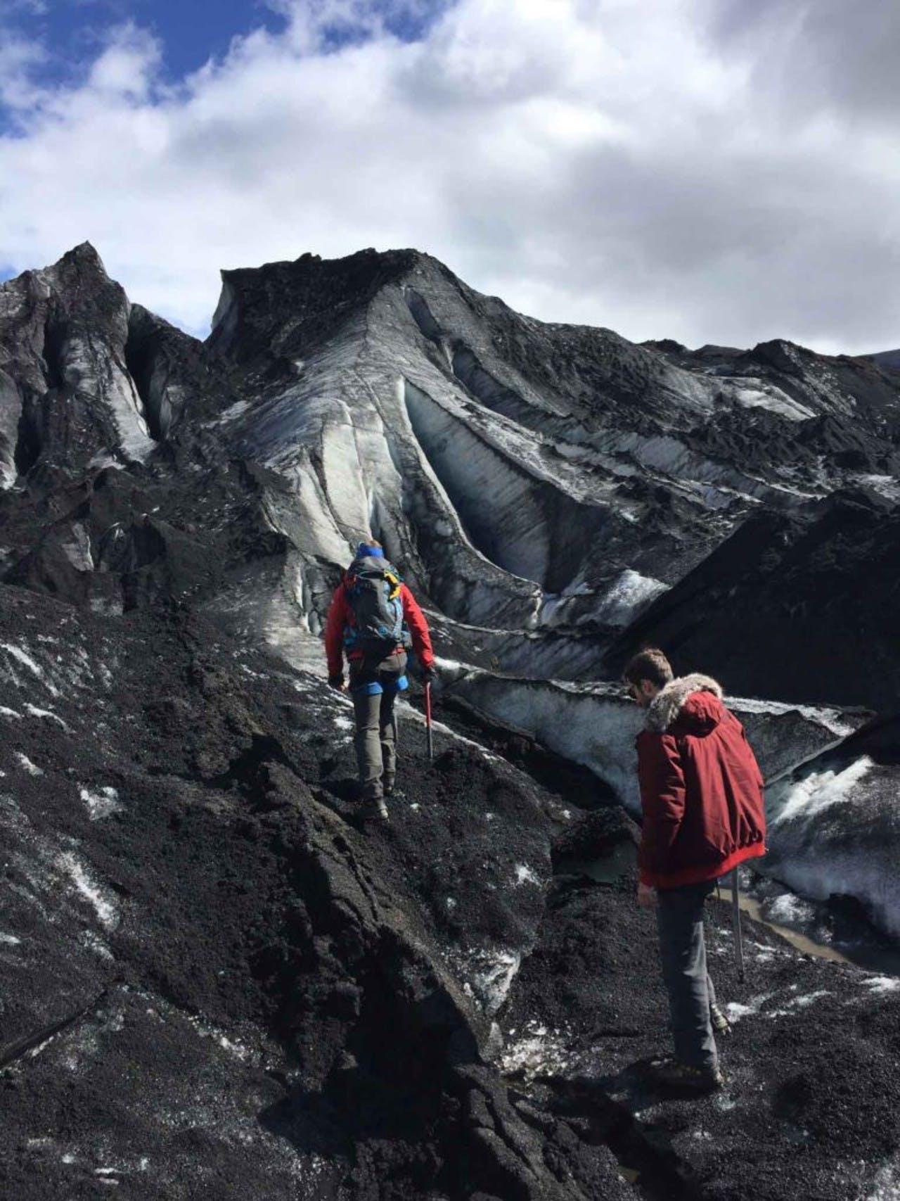 Two men hiking up a glacier covered in black sand