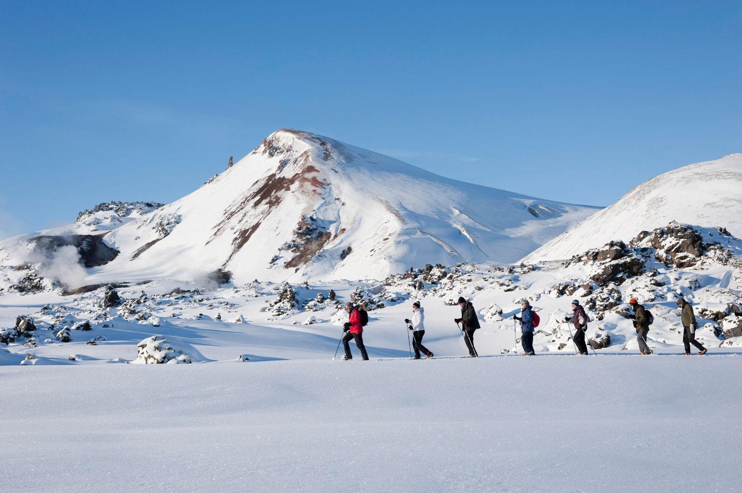 A group of hikers is shown walking in a line across a snowy field with a mountain in the background under a clear blue sky.