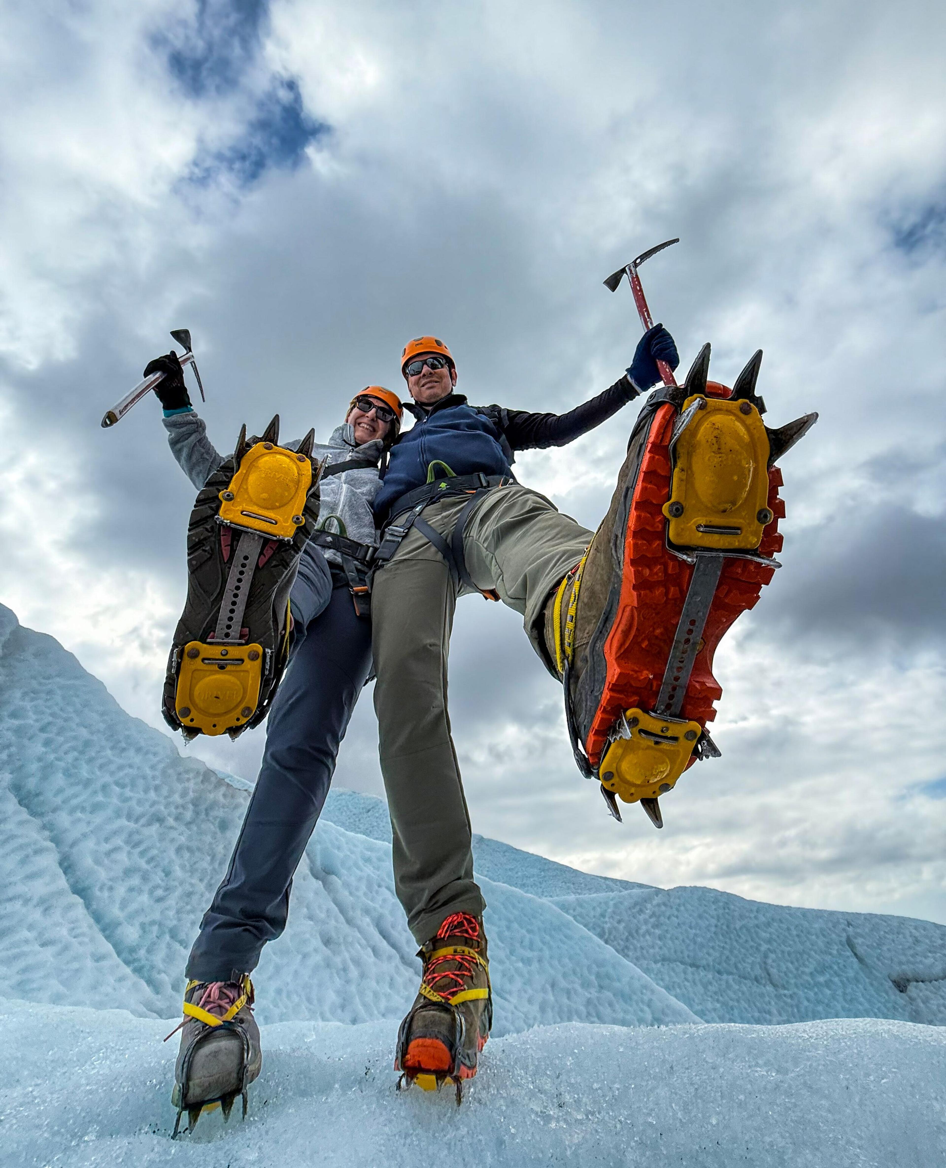 Two glacier hikers playfully raise their crampon-fitted boots and ice axes toward the camera, showcasing their gear against a cloudy sky
