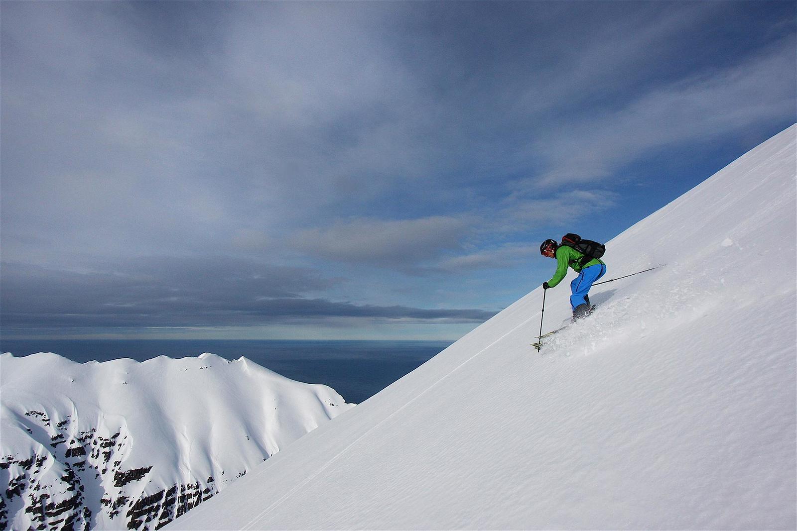 Skiier going down steep slaop and mountains across the fjord in the  background