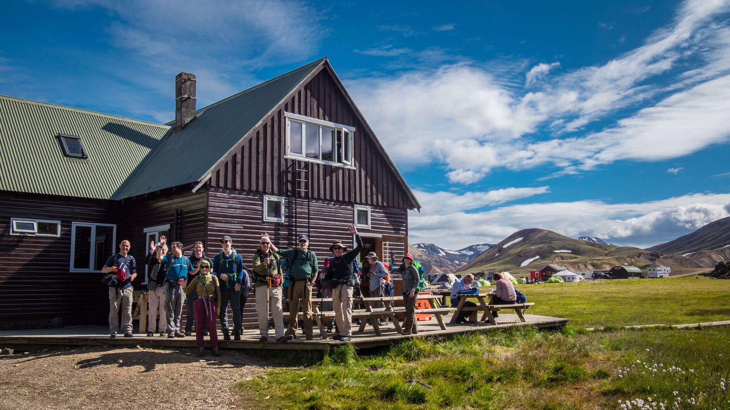 A group of hikers poses in front of a wooden lodge at Landmannalaugar base camp, with colorful mountains and tents visible in the background.
