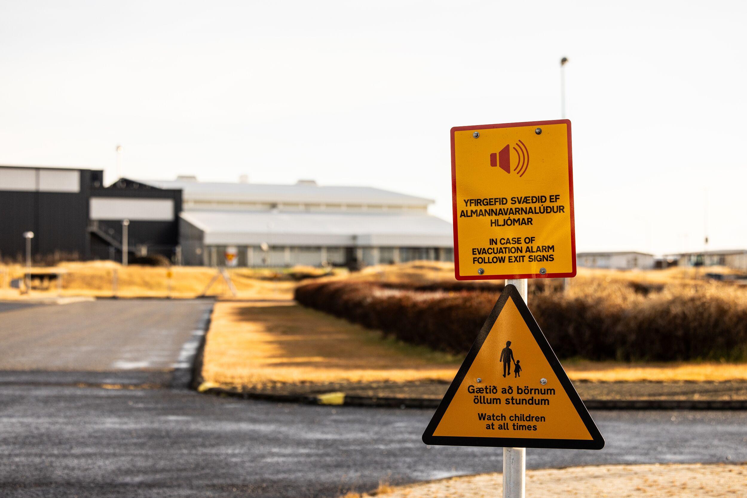 Bright yellow signs warn of evacuation procedures and safety precautions near an industrial building in Grindavík, highlighting the area's volcanic risk on the Reykjanes Peninsula. The signs read “In case of evacuation, follow exit signs” and “Watch children at all times.