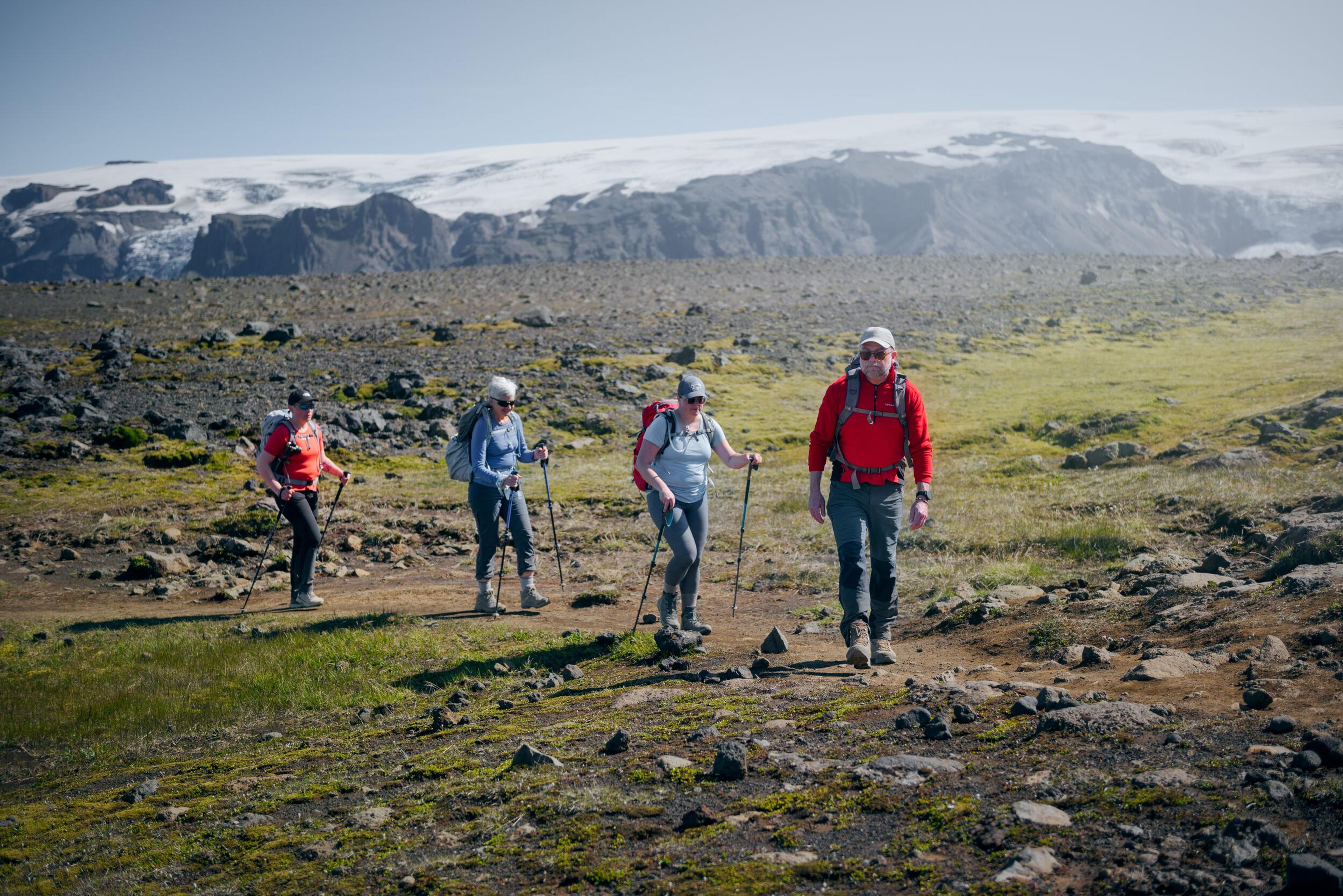 Hikers walking along a rocky trail in Landmannalaugar, with a glacier and lava field in the distance.