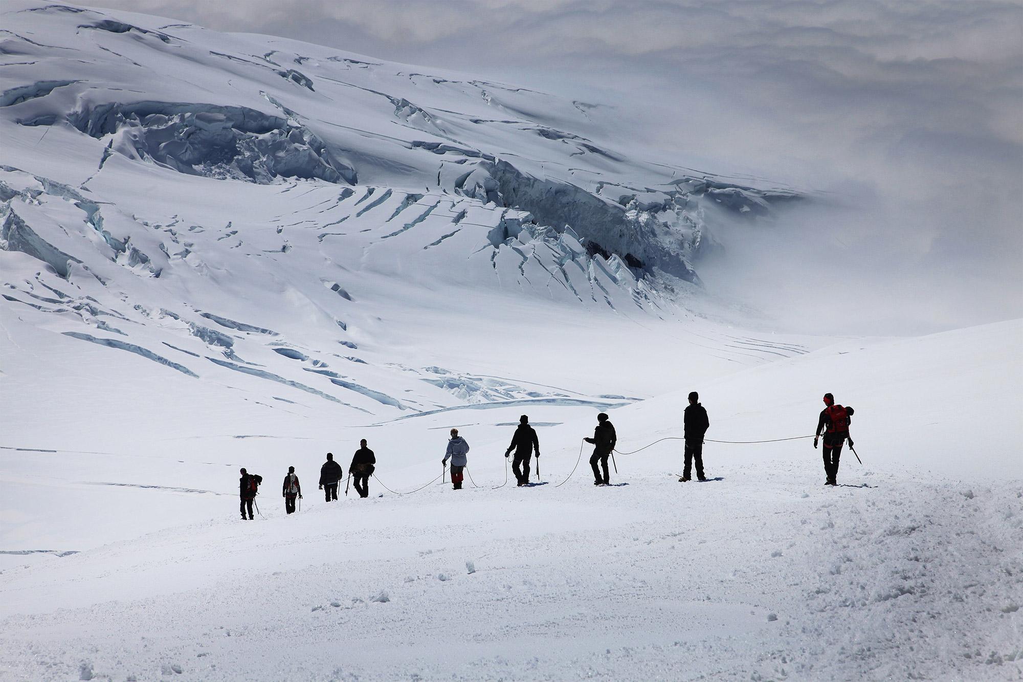 A group of mountaineers walking down Hvannadalshnúkur with bluish white mountains in the distance.