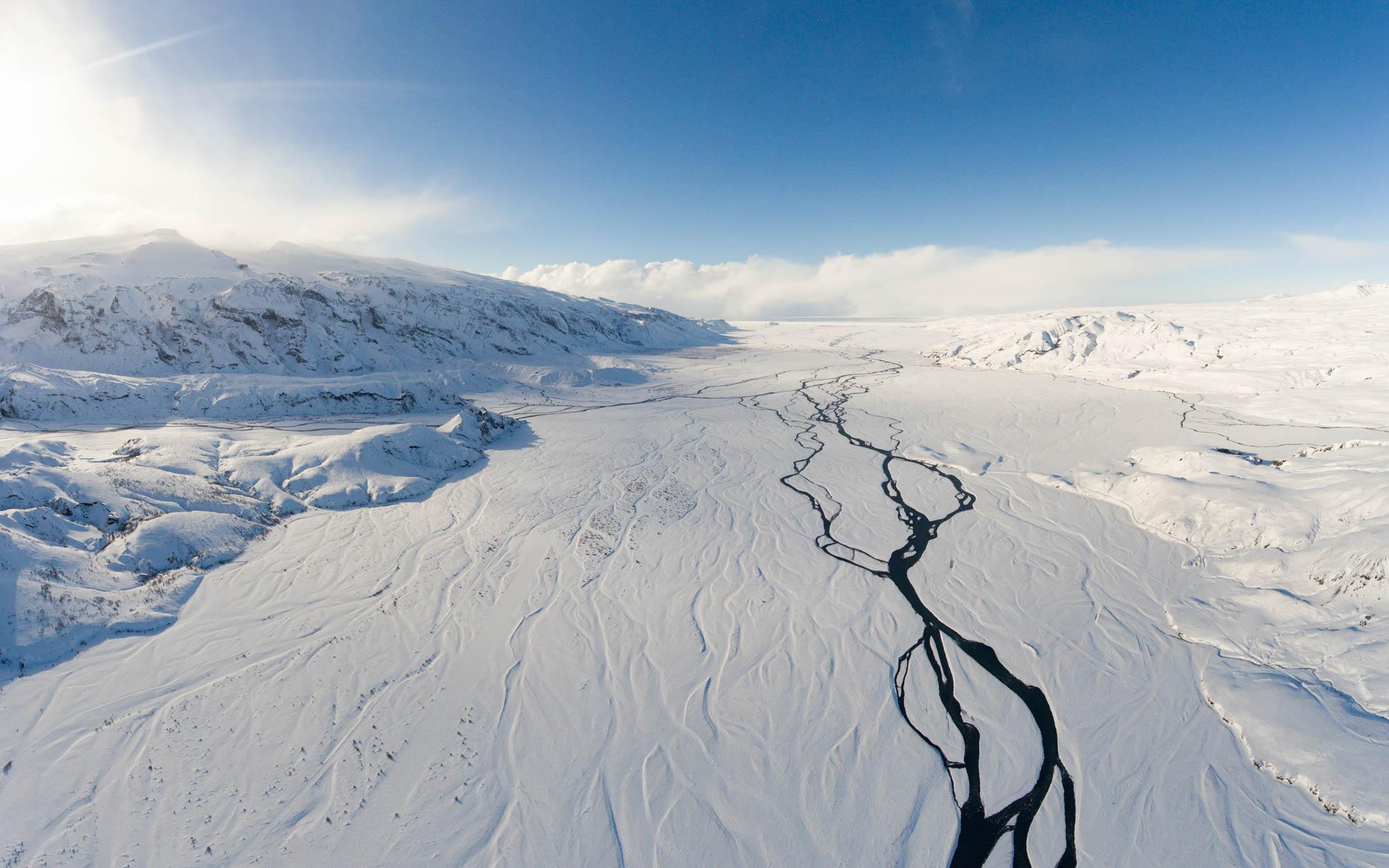 An aerial view of a snowy landscape featuring a meandering black river cutting through the white terrain under a clear blue sky.
