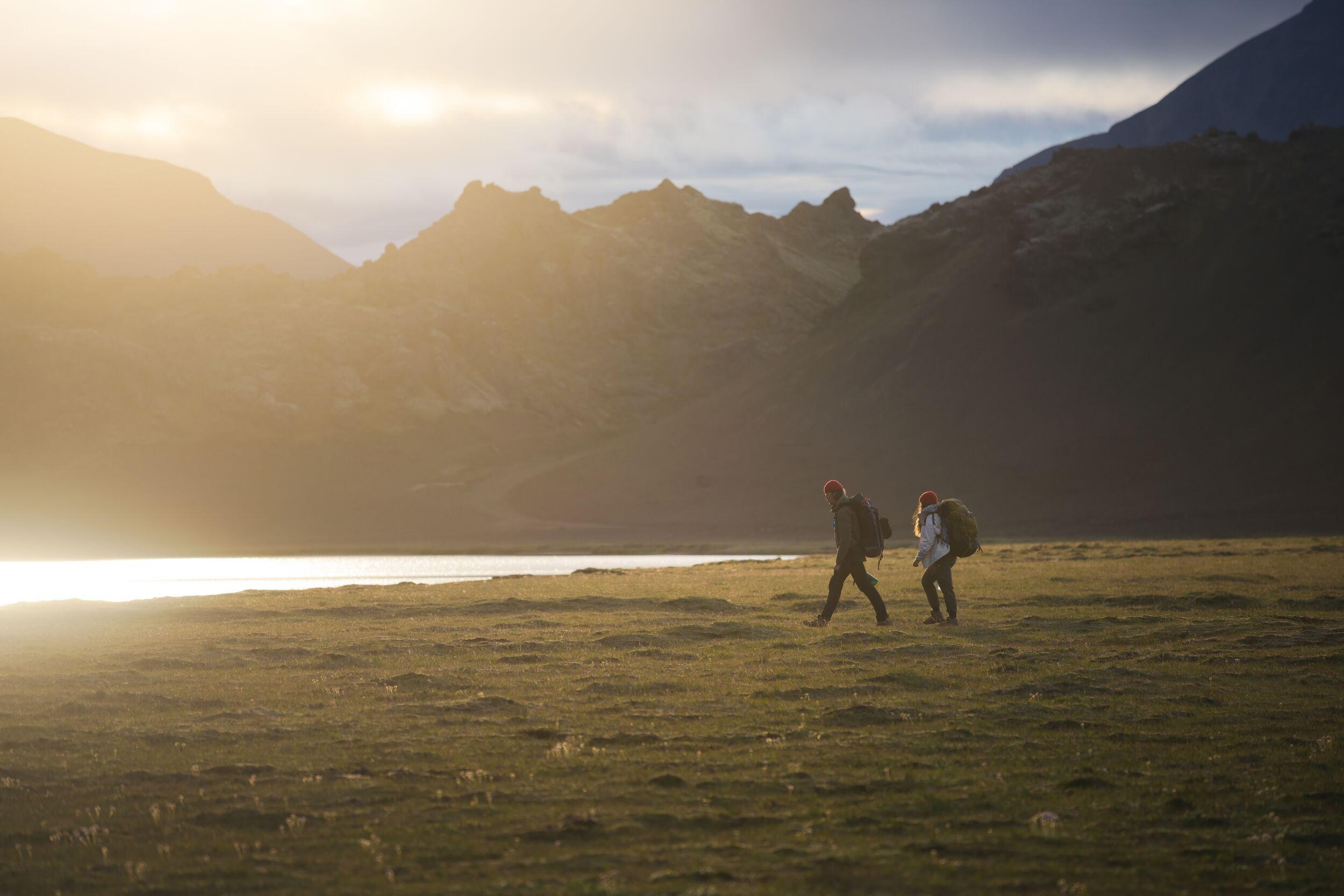2 hikers walk on grass with mountains in the background and the shining sun