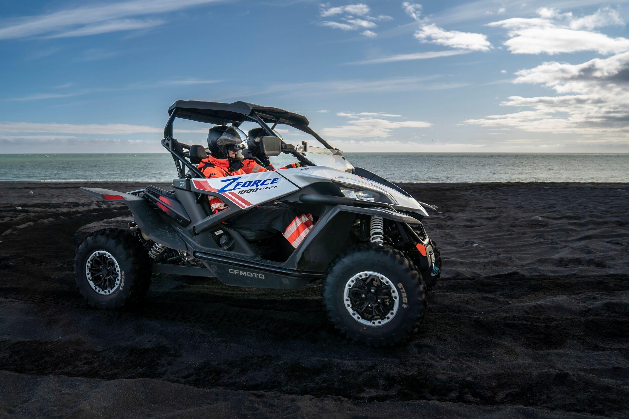 A rugged off-road buggy with "Z Force" branding is parked on a black sand beach with the ocean and blue sky in the background. A driver in bright orange gear and a helmet sits inside, ready for an Icelandic South Coast adventure.