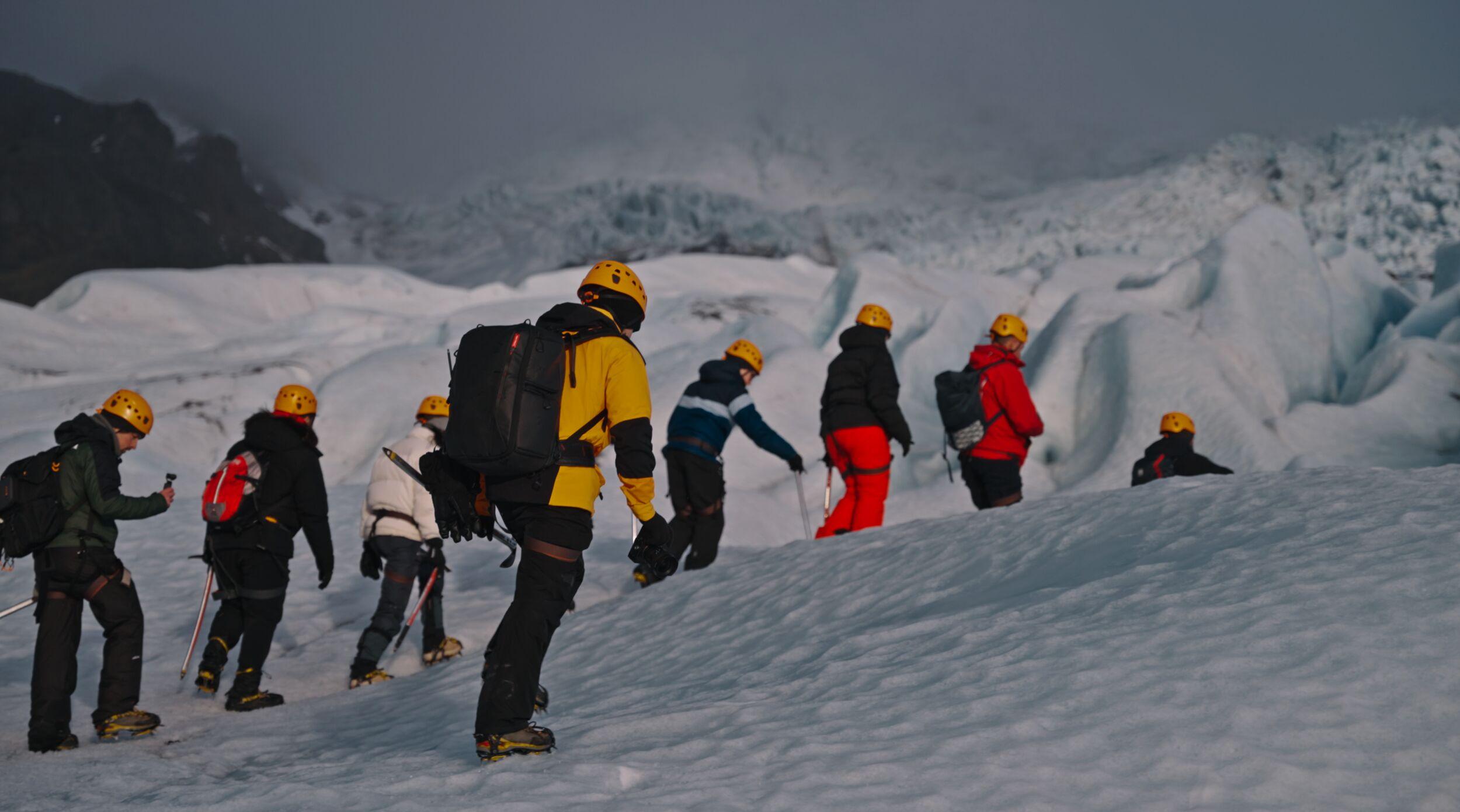 Group of people wearing helmets and crampons hiking up an icy slope on a glacier in Iceland, surrounded by snow and dramatic terrain.