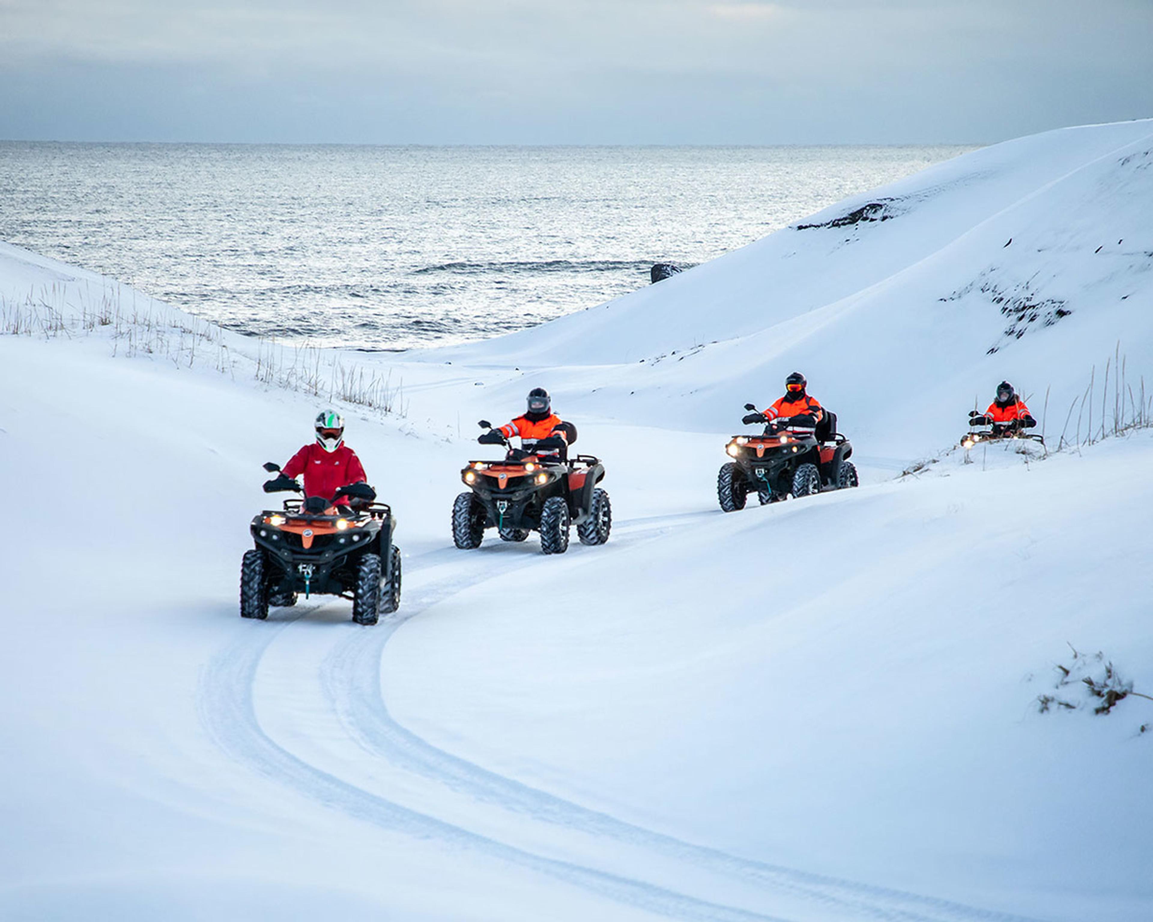 ATV quad riders driving between snow covered sand dunes and the ocean in the background