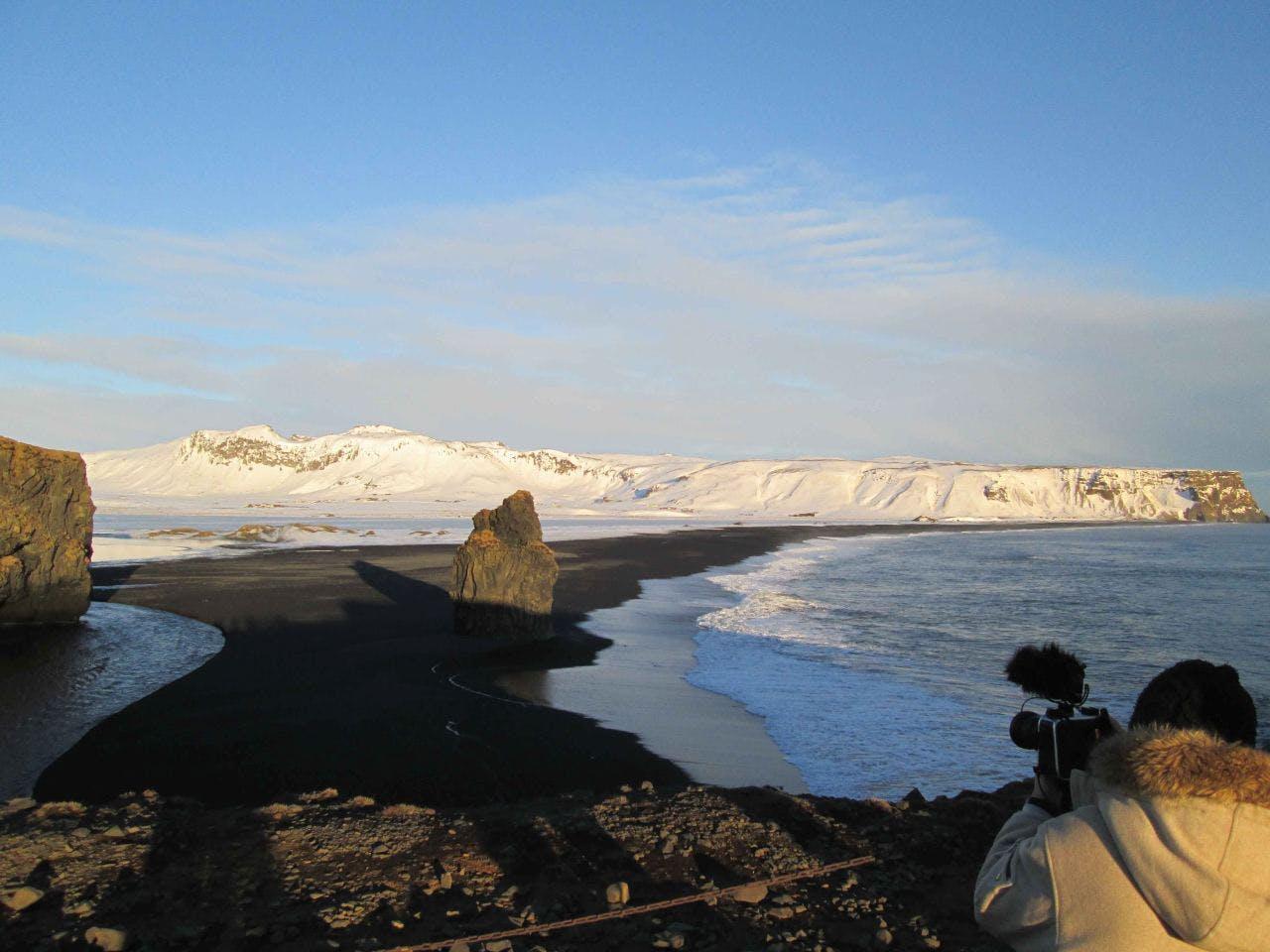 man photographing a big rock on a black beach with snowy mountains and blue sky in the background