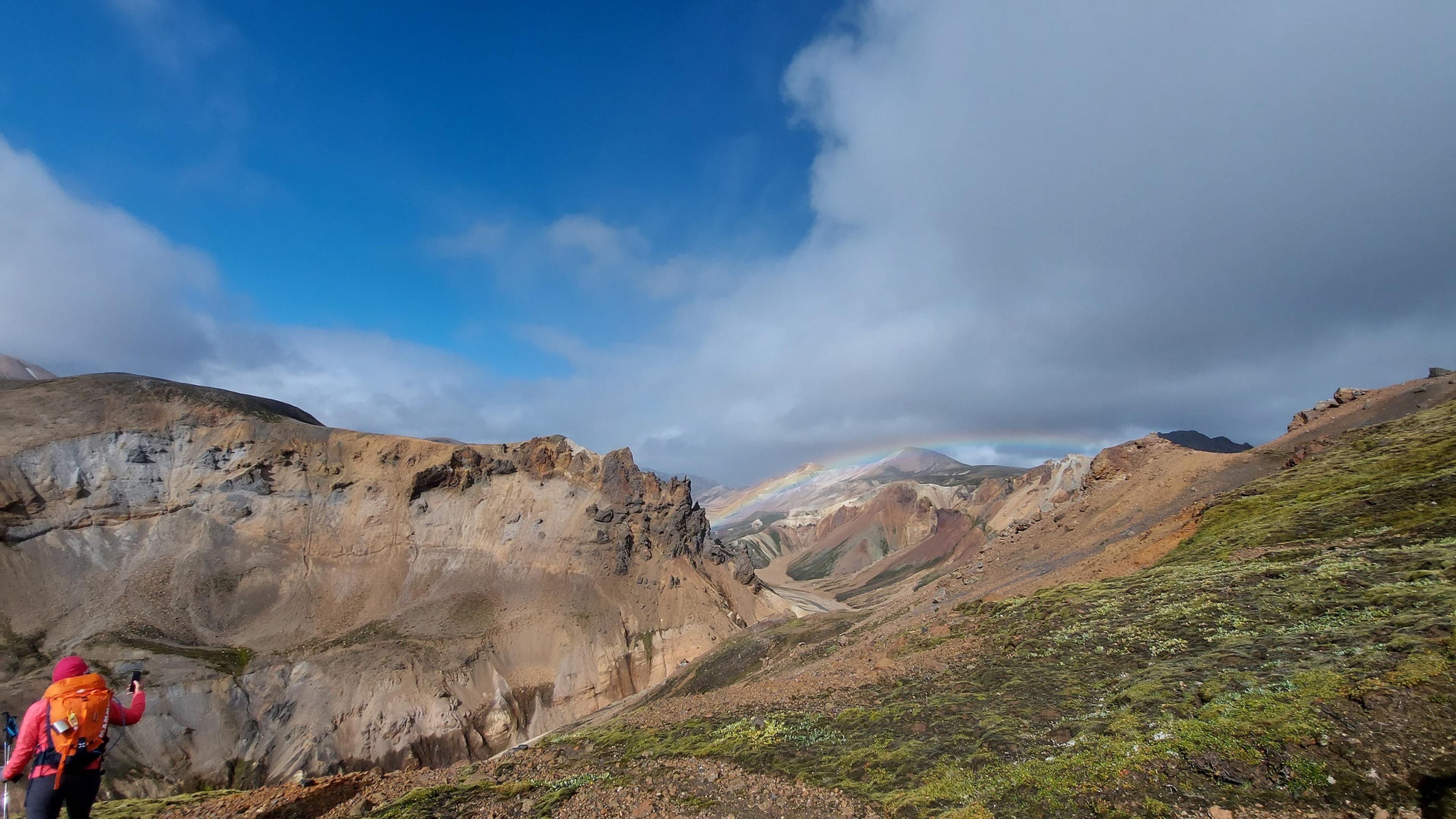 A hiker with a bright orange backpack traverses a grassy ridge in Landmannalaugar, overlooking colorful rhyolite mountains and a faint rainbow under a cloudy sky.