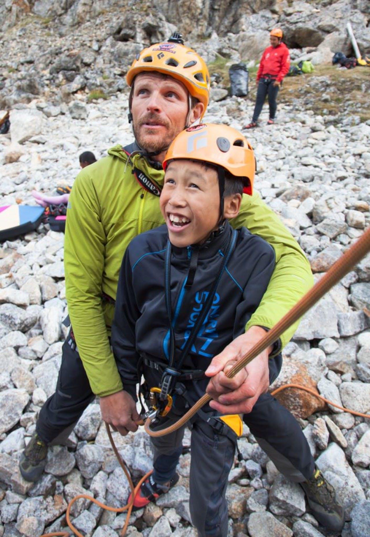 a boy and a guide in a yellow jacket getting read for rock climbing while a man in a red jacket is in the background