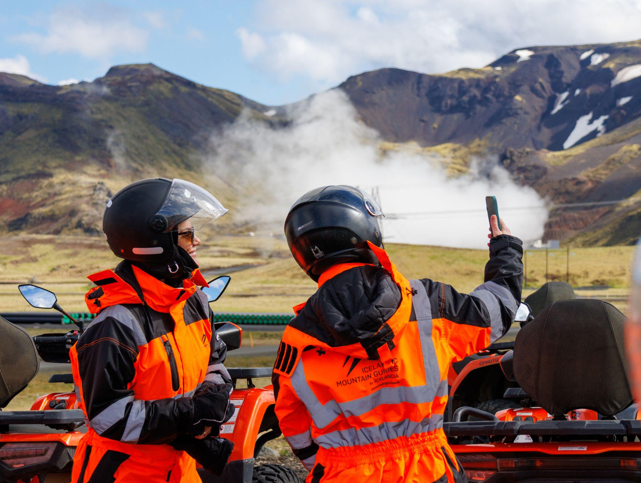 Two people standing beside their ATV, taking a selfie with geothermal steam in the background.