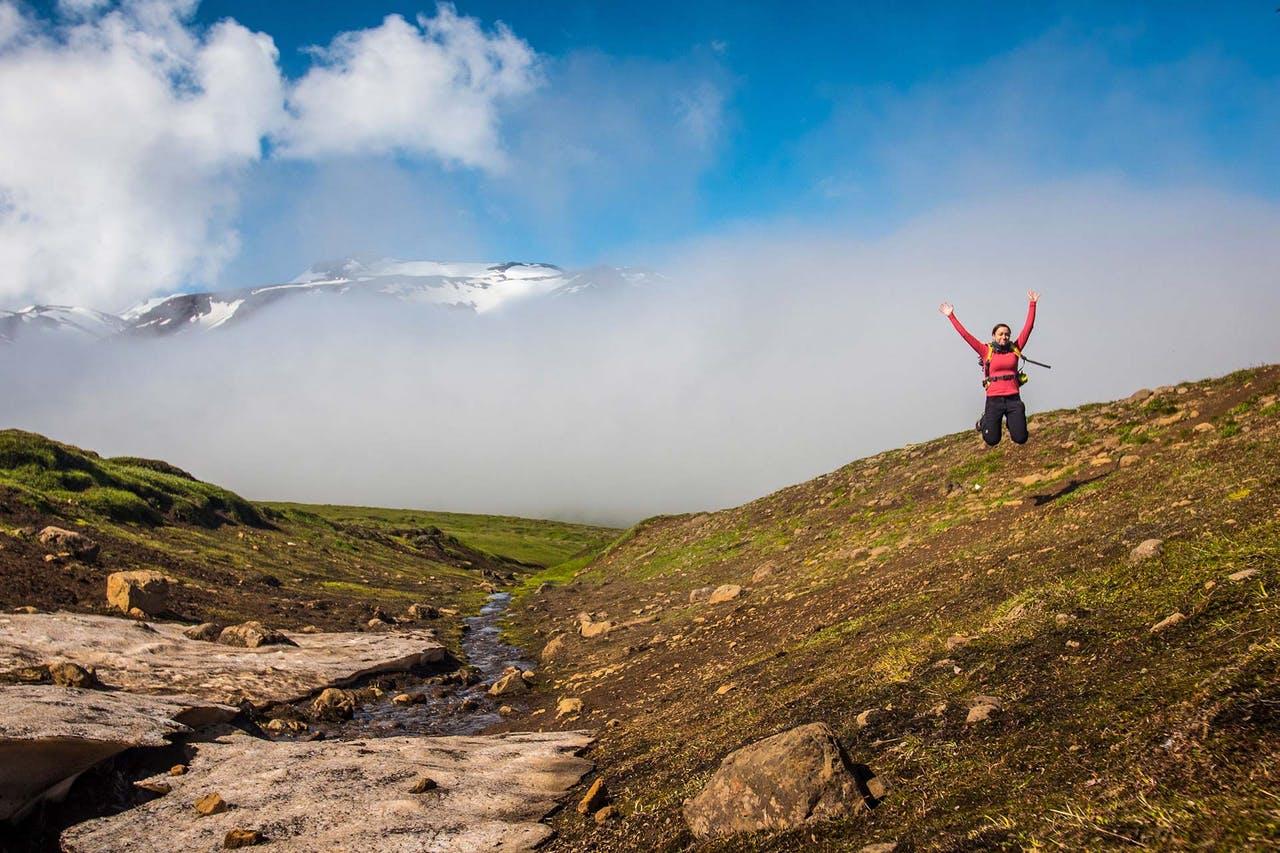 Woman in red on a hike in Iceland jumping in nature on a green hill with semi-cloudy blue sky in the background