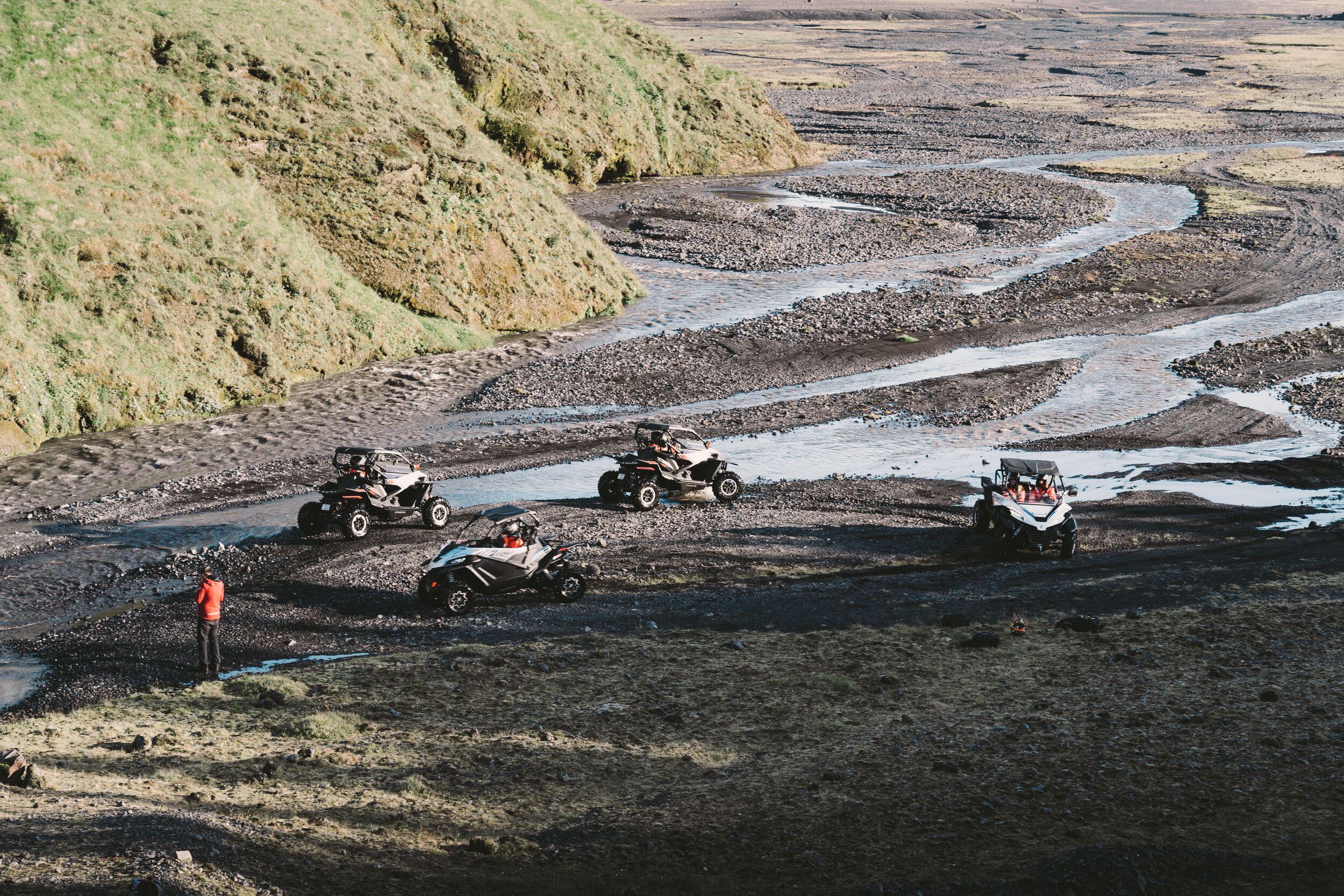 Several off-road buggies are parked by a braided riverbed in a scenic valley, with a guide standing nearby in bright gear.