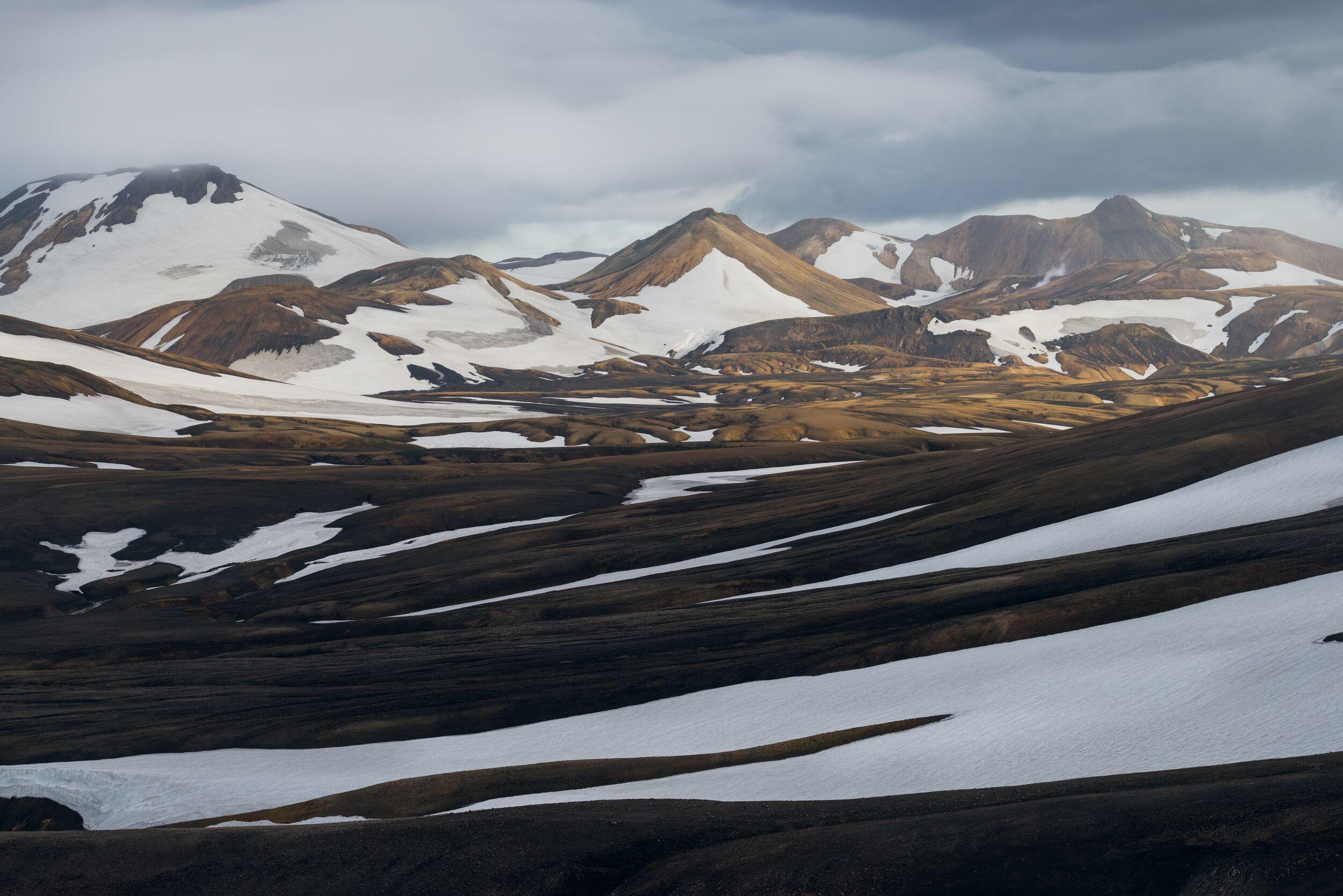 A view of Landmannalaugar’s volcanic landscape with snow-covered slopes, dark lava fields, and distant rugged mountains under an overcast sky.