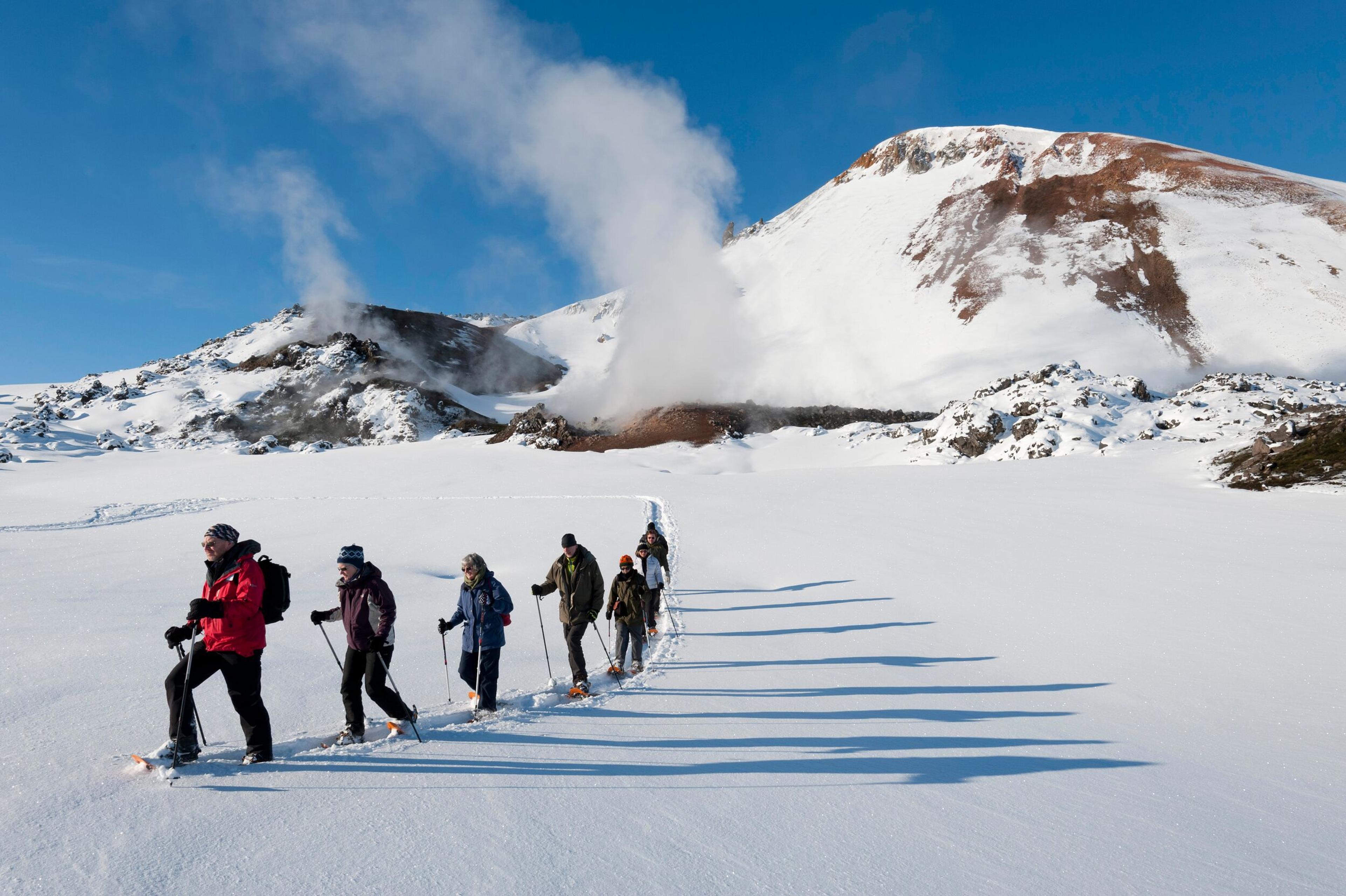 A line of hikers in winter gear traversing a snow-covered landscape in Landmannalaugar, passing by steaming geothermal vents and volcanic peaks.