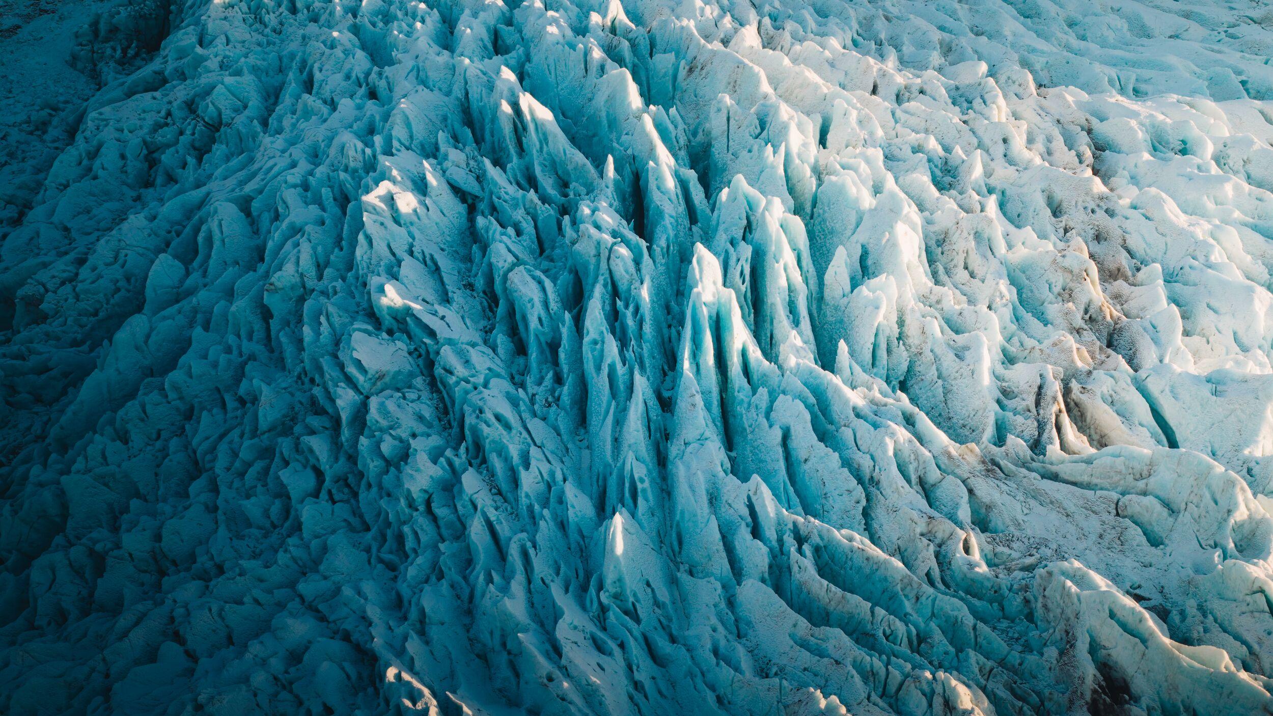Aerial close-up of sharp, jagged ice pinnacles and seracs on a glacier, glowing blue and white in natural light