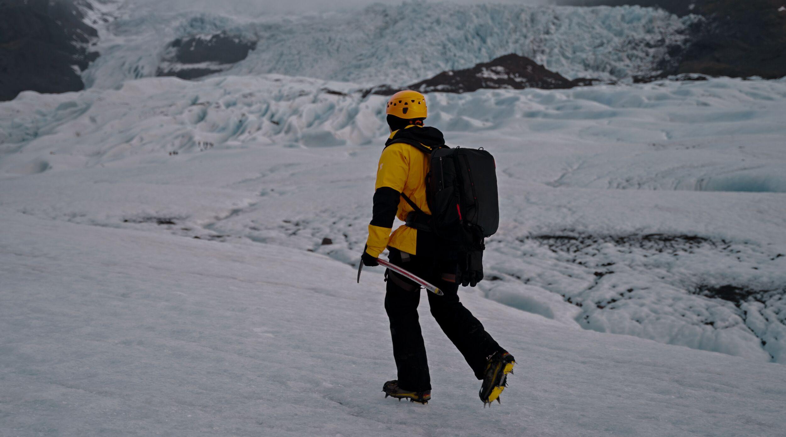 Person wearing a yellow jacket and helmet walking with crampons and an ice axe on a glacier in Iceland, surrounded by dramatic ice formations and snowy terrain.        You said: