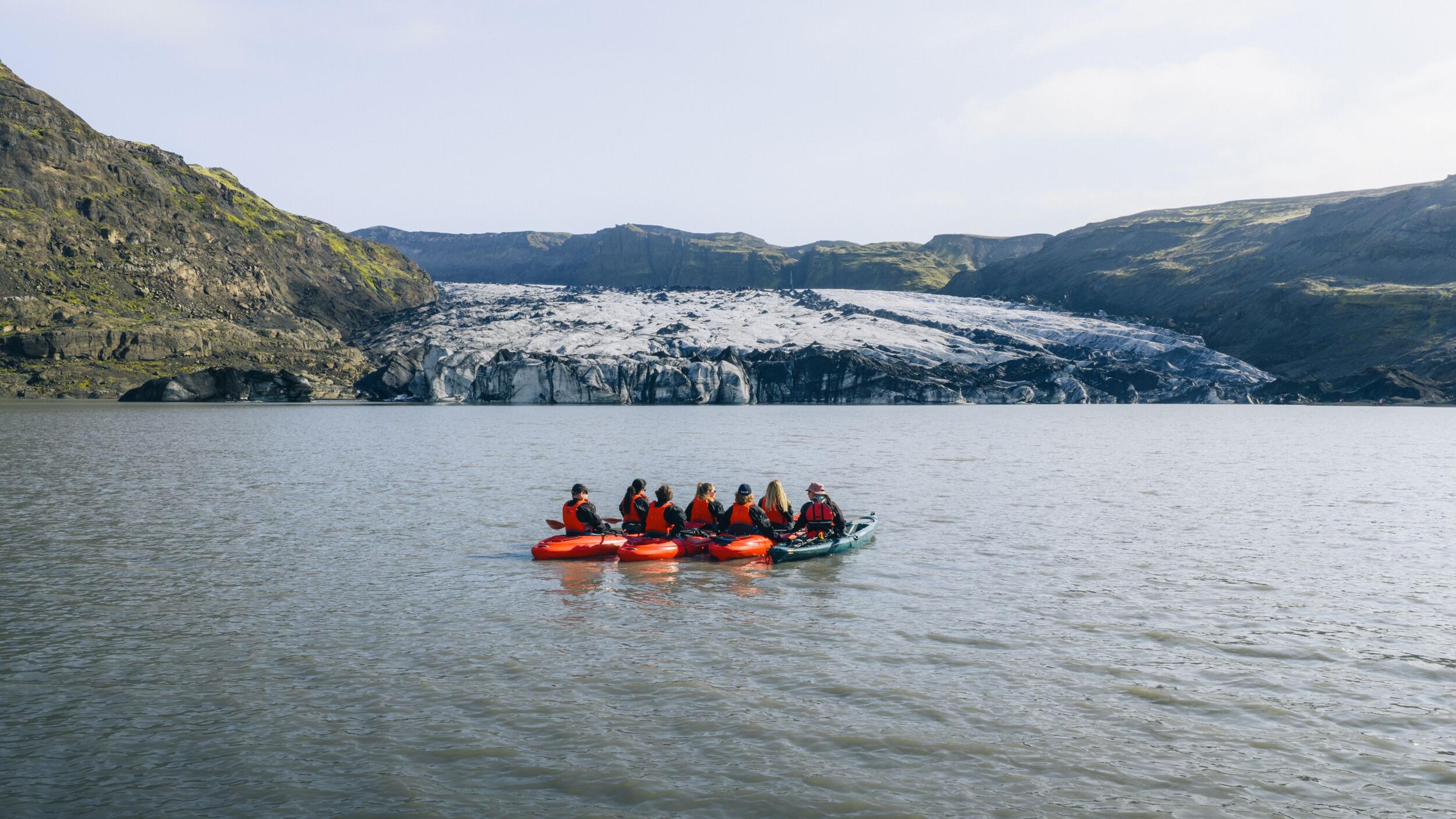 A group of people in orange life jackets paddles inflatable kayaks across a glacial lake, with a large glacier and rugged, moss-covered mountains in the background.
