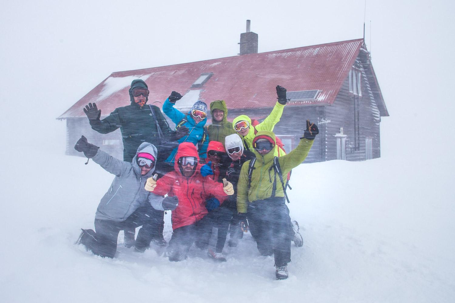 A group of people in colorful winter clothing are posing joyfully in front of a snow-covered cabin, with some individuals raising their arms in celebration.