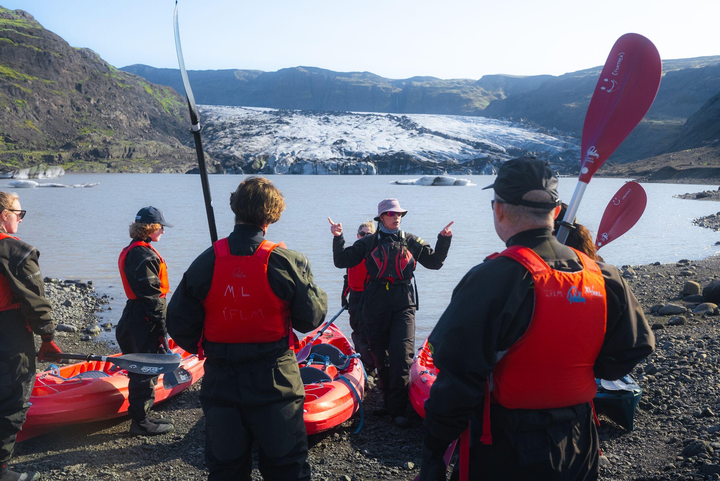 A group of people wearing red life jackets and carrying paddles prepare inflatable kayaks on a rocky lakeshore, with a large glacier and rugged mountains in the background.