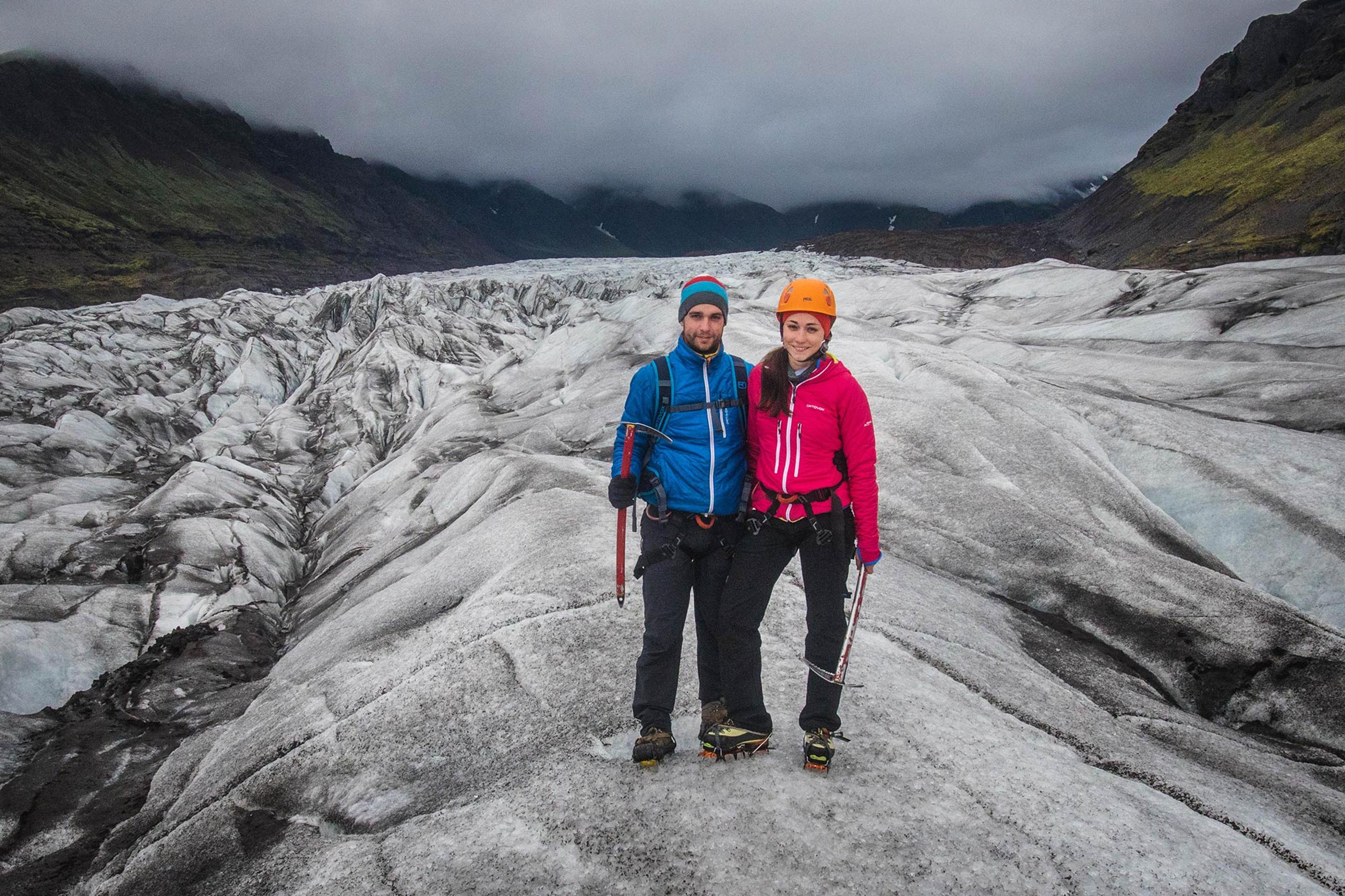 A couple, dressed in colorful outdoor gear and crampons, stands together on a glacier with a dramatic, cloudy mountain backdrop.