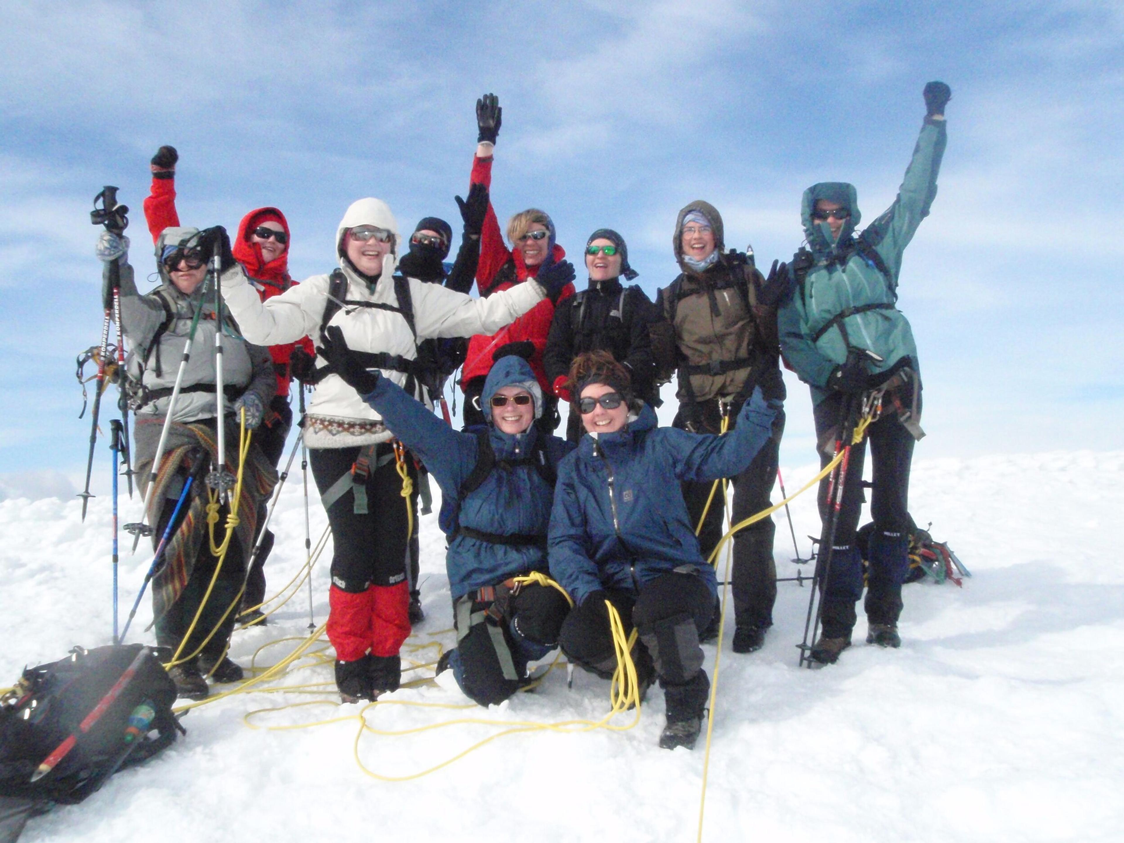 A jubilant group of hikers in winter gear celebrates on the snowy summit of Eyjafjallajökull, with arms raised and smiles, against a backdrop of a clear sky.