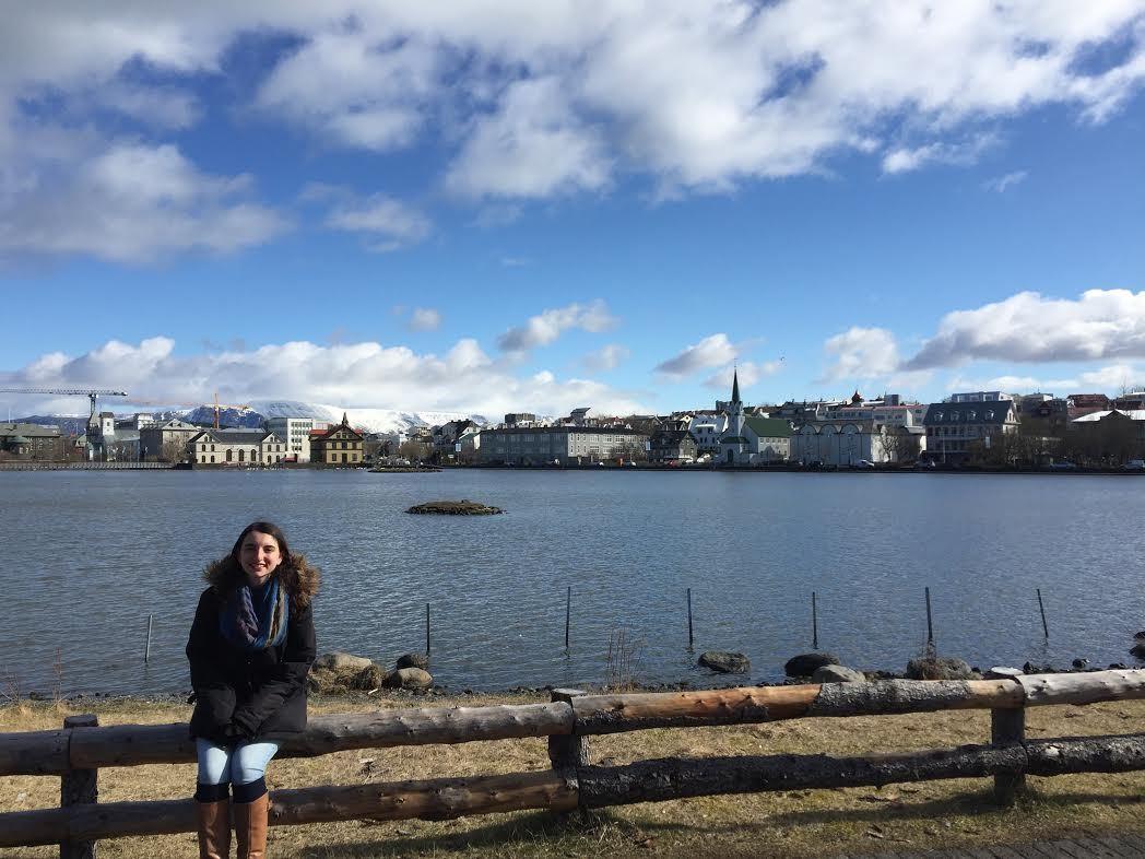 Sydney, Icelandic Mountain Guide's Adventurer of the Week, is sitting by the shore with a blue sky and buildings in the background.