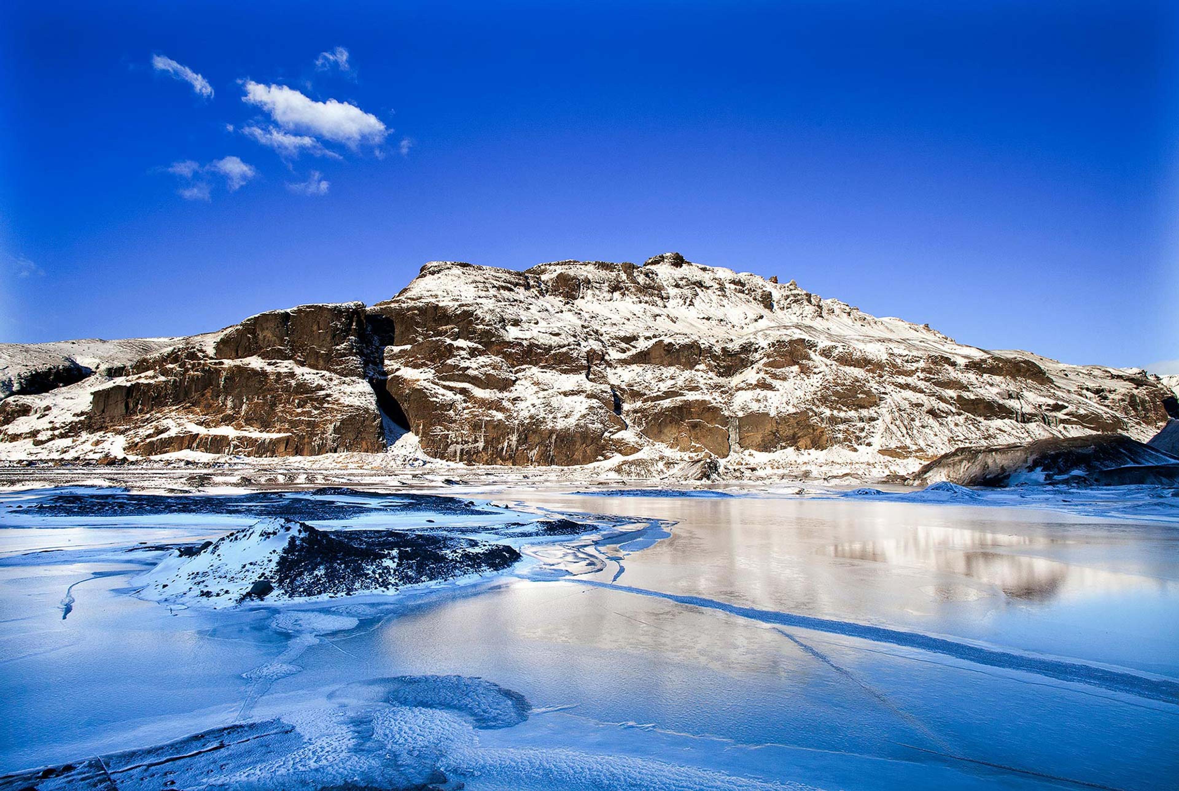 A brown mountain covered in snow behind frozen blue water and a bright blue sky on the South Coast in Iceland
