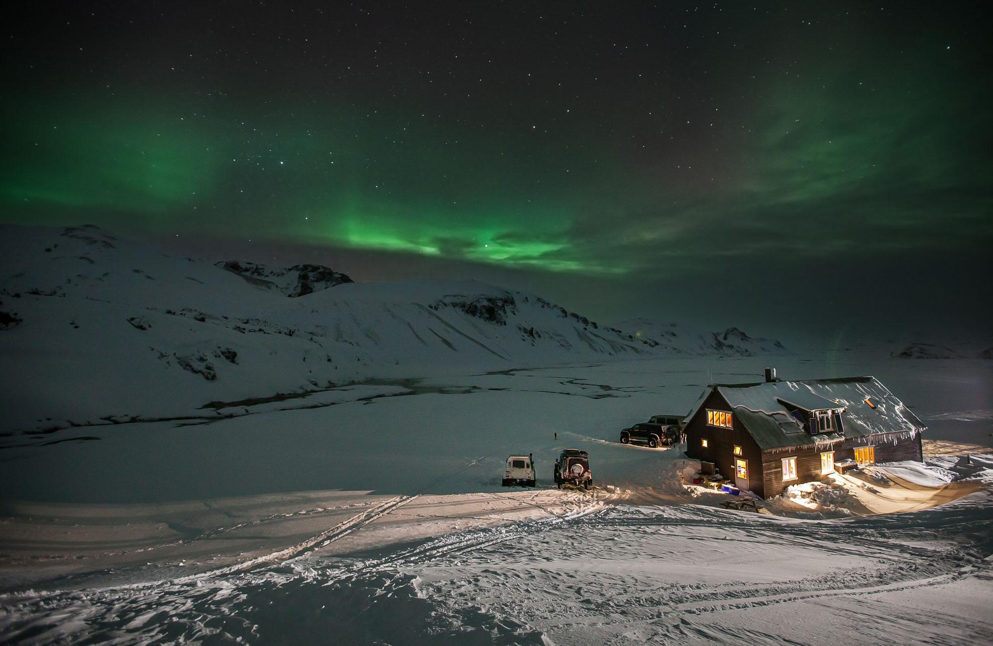 A cozy cabin illuminated from within is set against a night sky with the Northern Lights, surrounded by a snowy landscape and mountains.