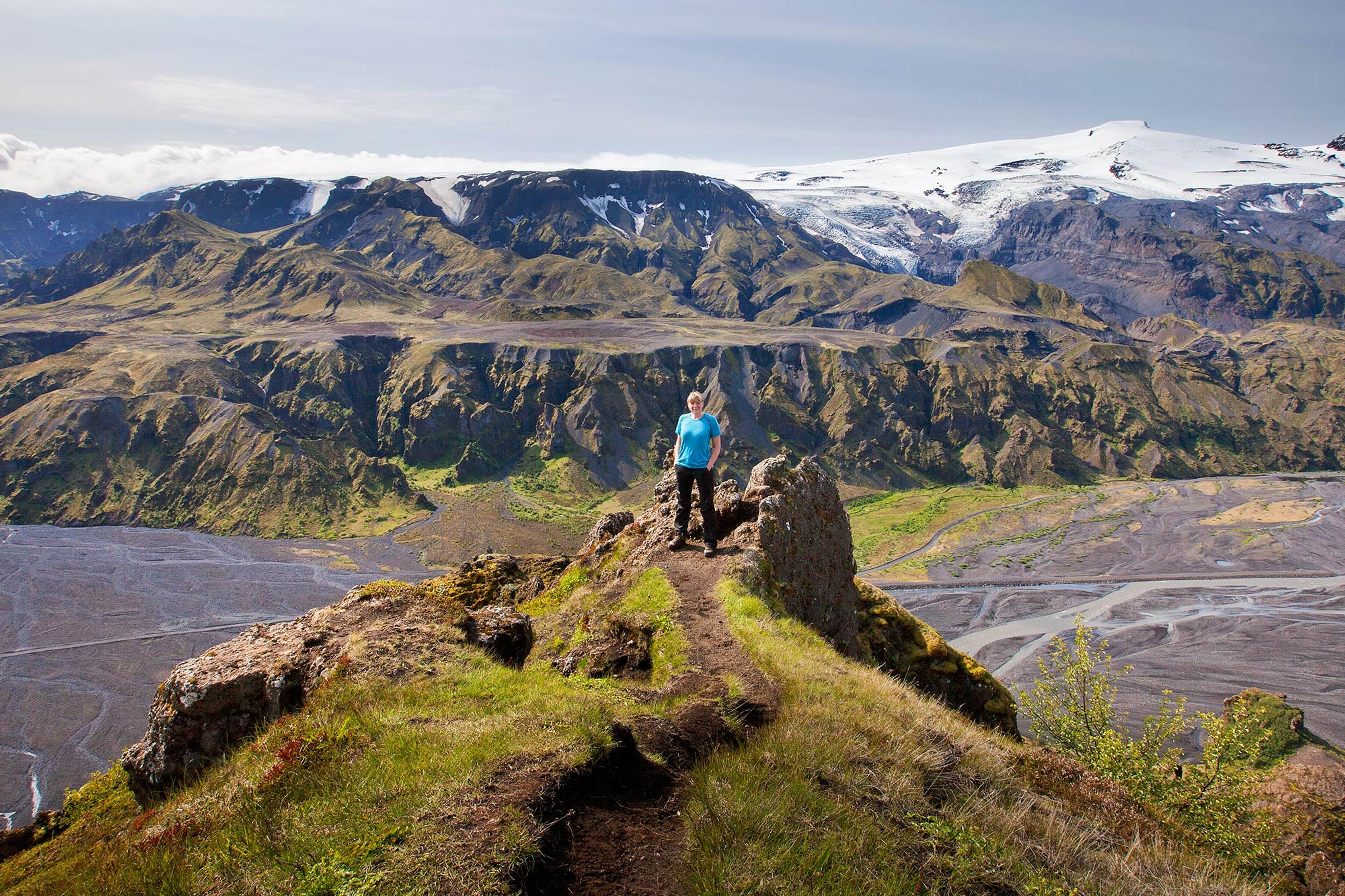 A hiker stands on a grassy ridge, overlooking a dramatic valley with rugged cliffs, braided riverbeds, and a glacier-capped mountain in the distance.