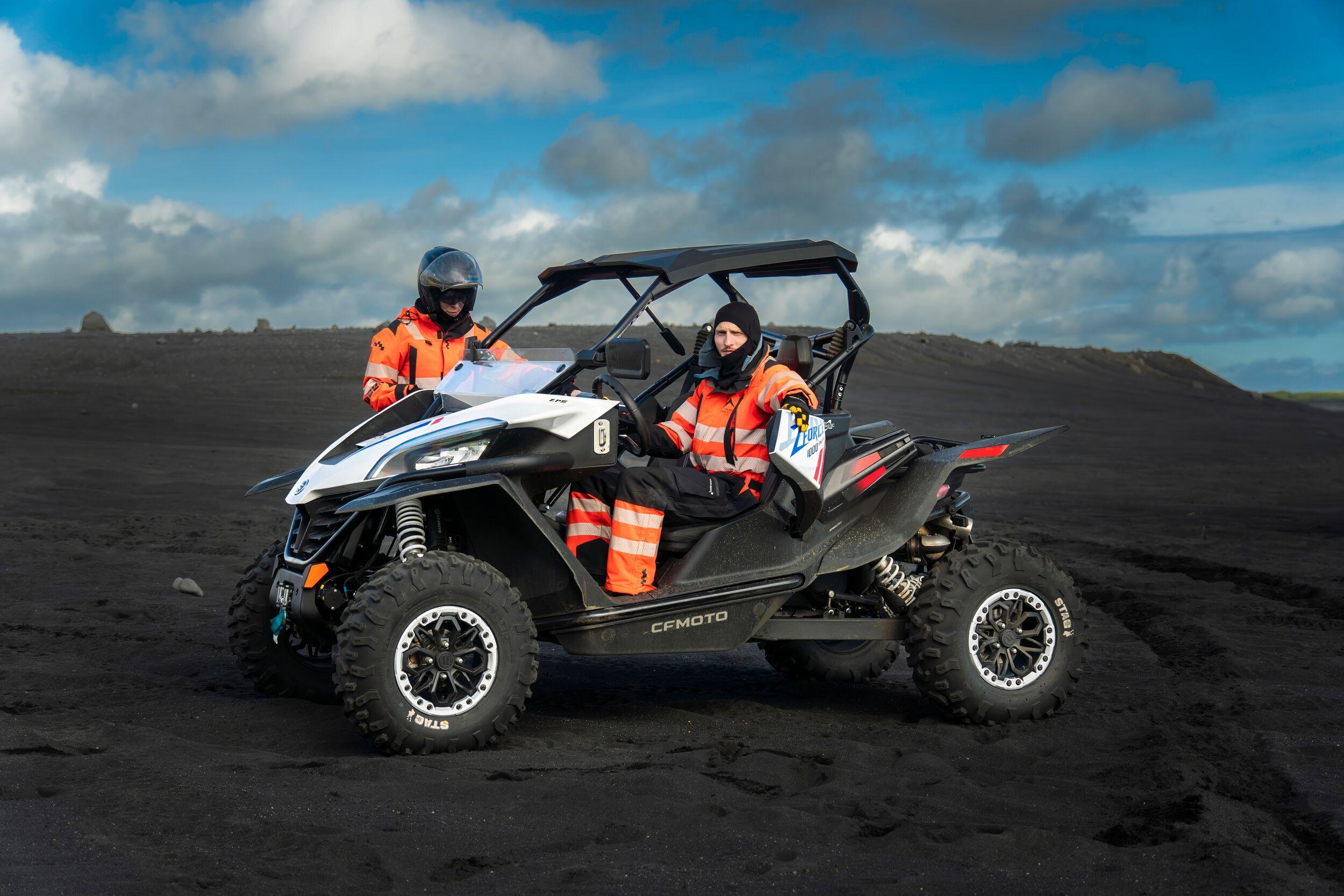 Two people in orange gear sitting in a black buggy on a vast black sand plain under a cloudy blue sky, ready for adventure.