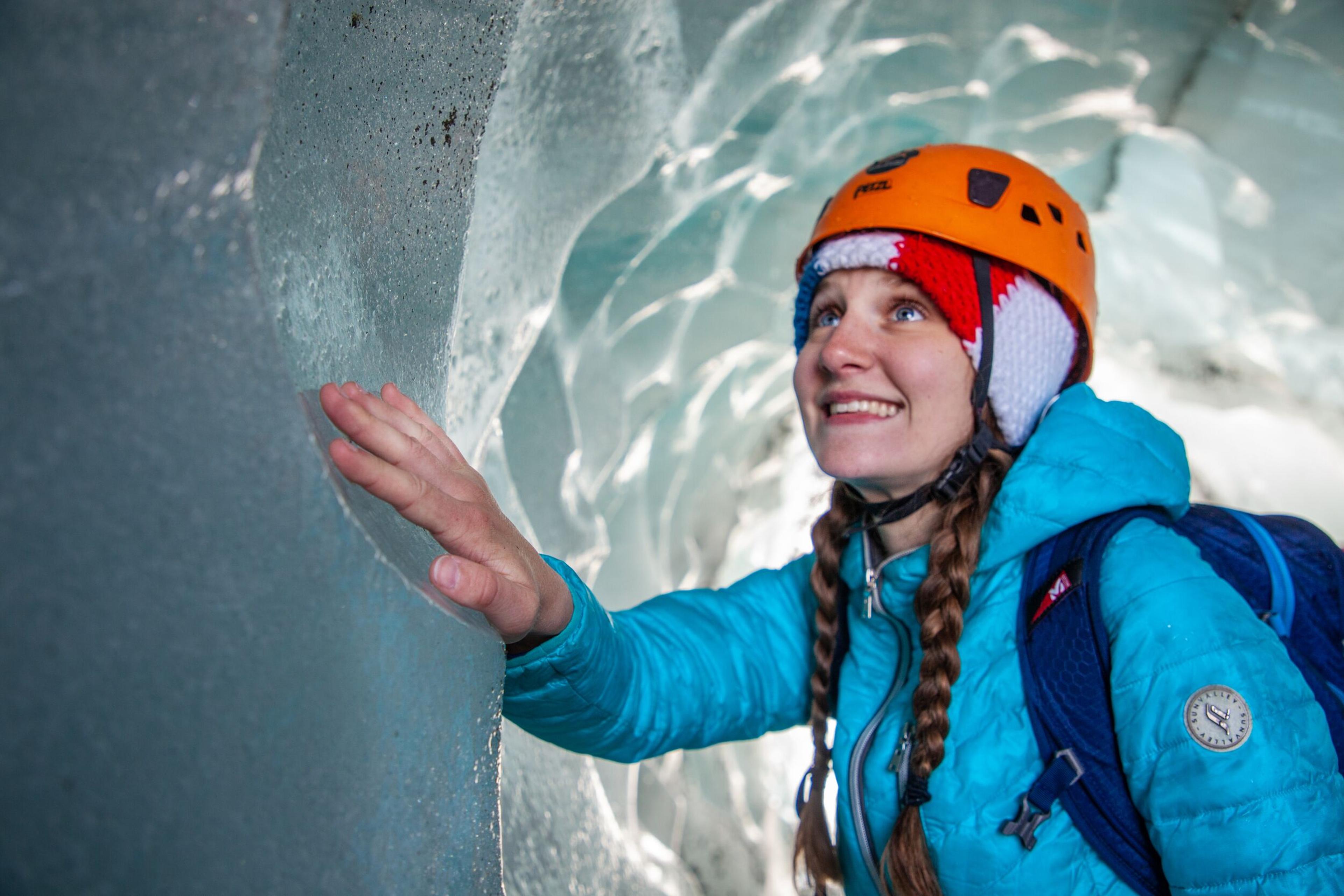 A person wearing orange helmet and blue jacket, touching the wall of an ice cave