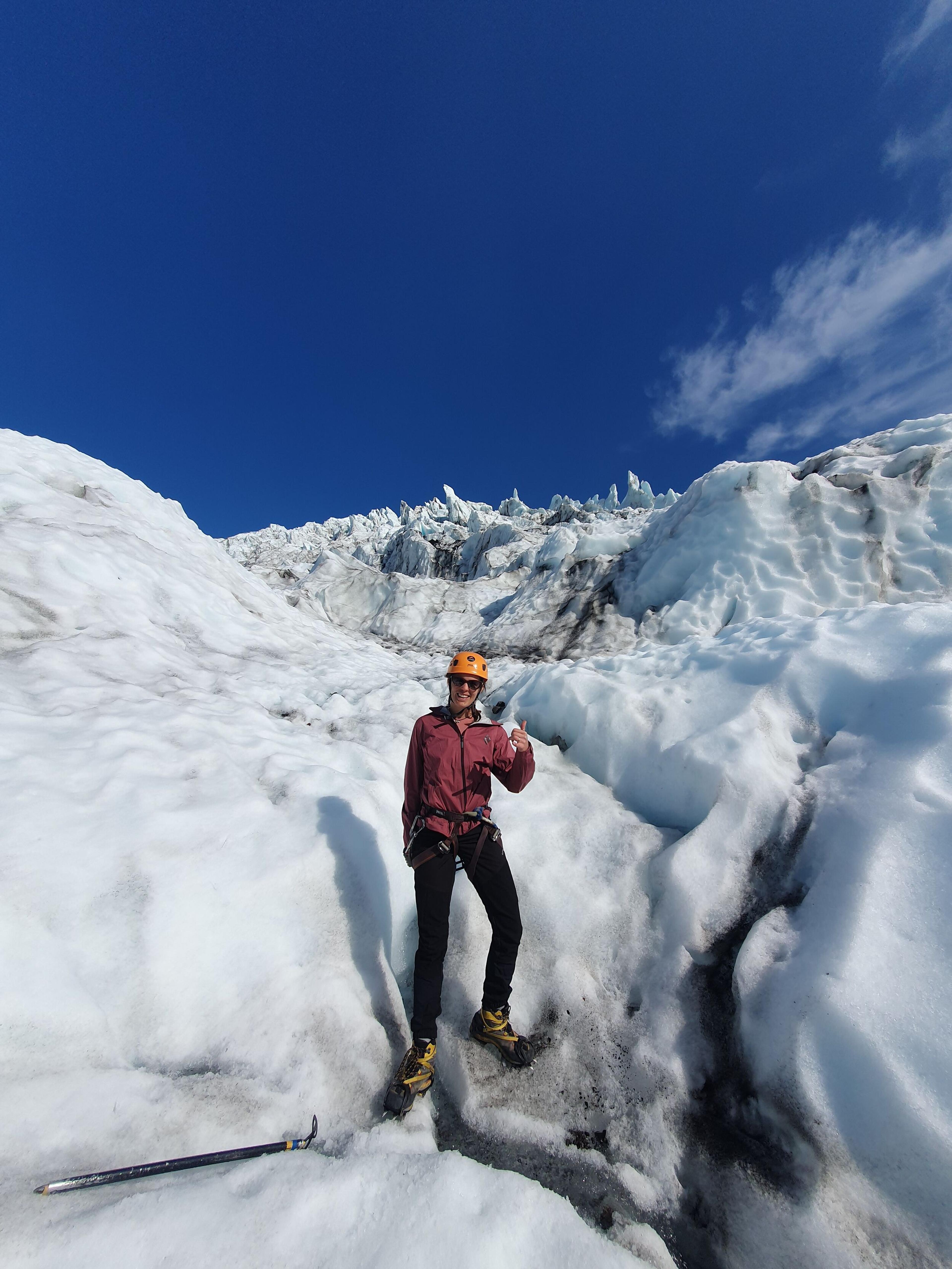 "A smiling hiker in a helmet and climbing harness gives a thumbs up while standing among towering blue-white ice formations on a glacier