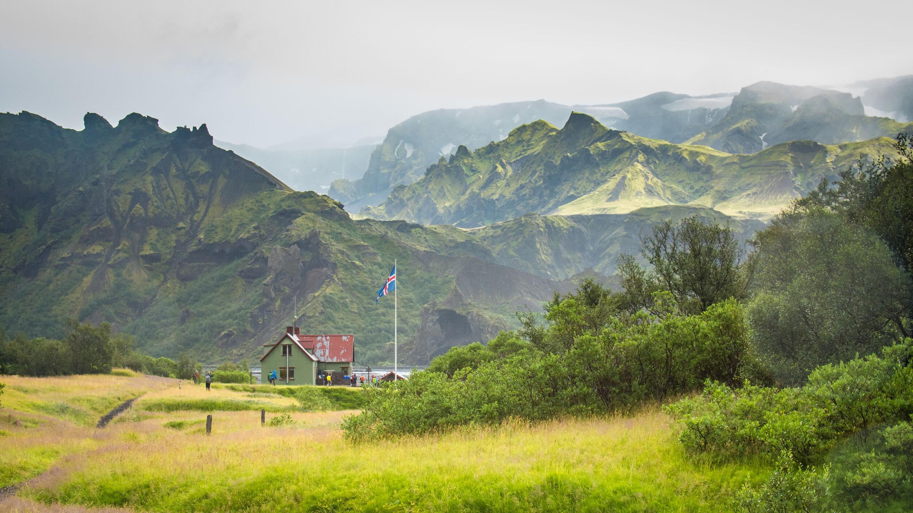 A small house with a red roof and an Icelandic flag stands in a lush green valley, surrounded by towering moss-covered mountains in the Icelandic Highlands. The misty atmosphere adds to the serene and picturesque landscape.