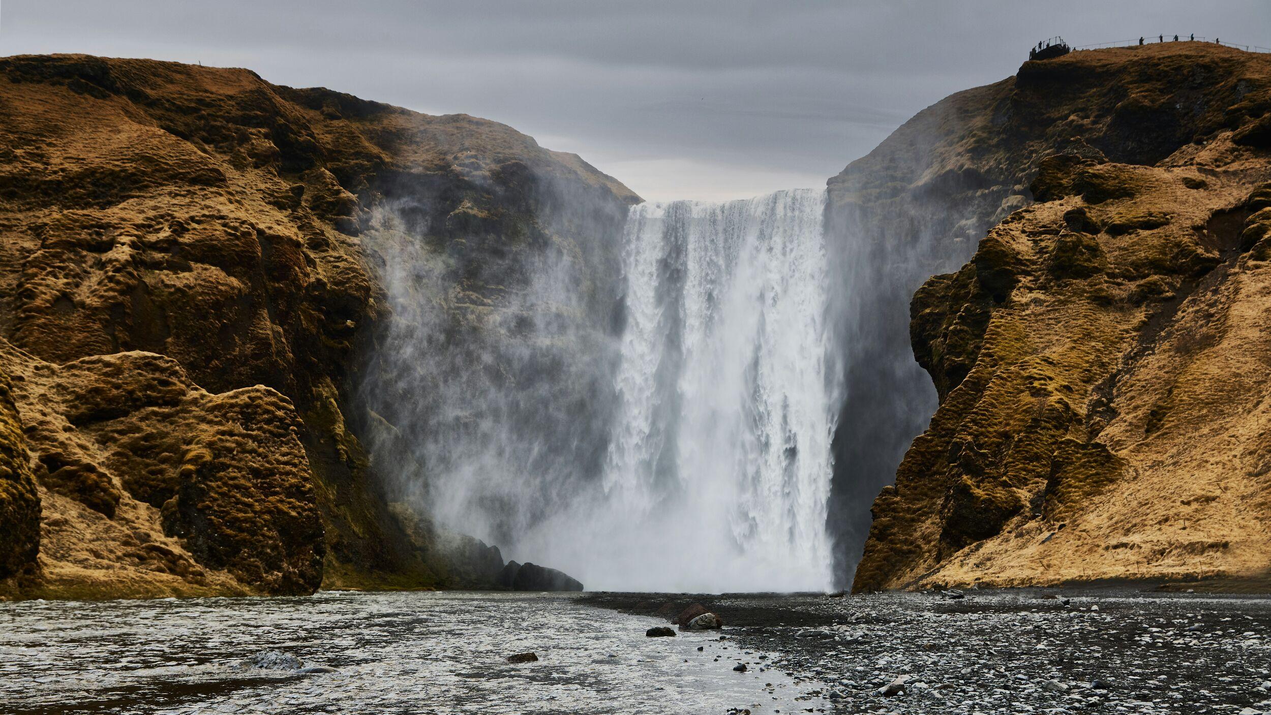 A powerful view of Skógafoss waterfall in Iceland, with water cascading down a steep cliff surrounded by rugged, moss-covered rock formations under a gray sky.