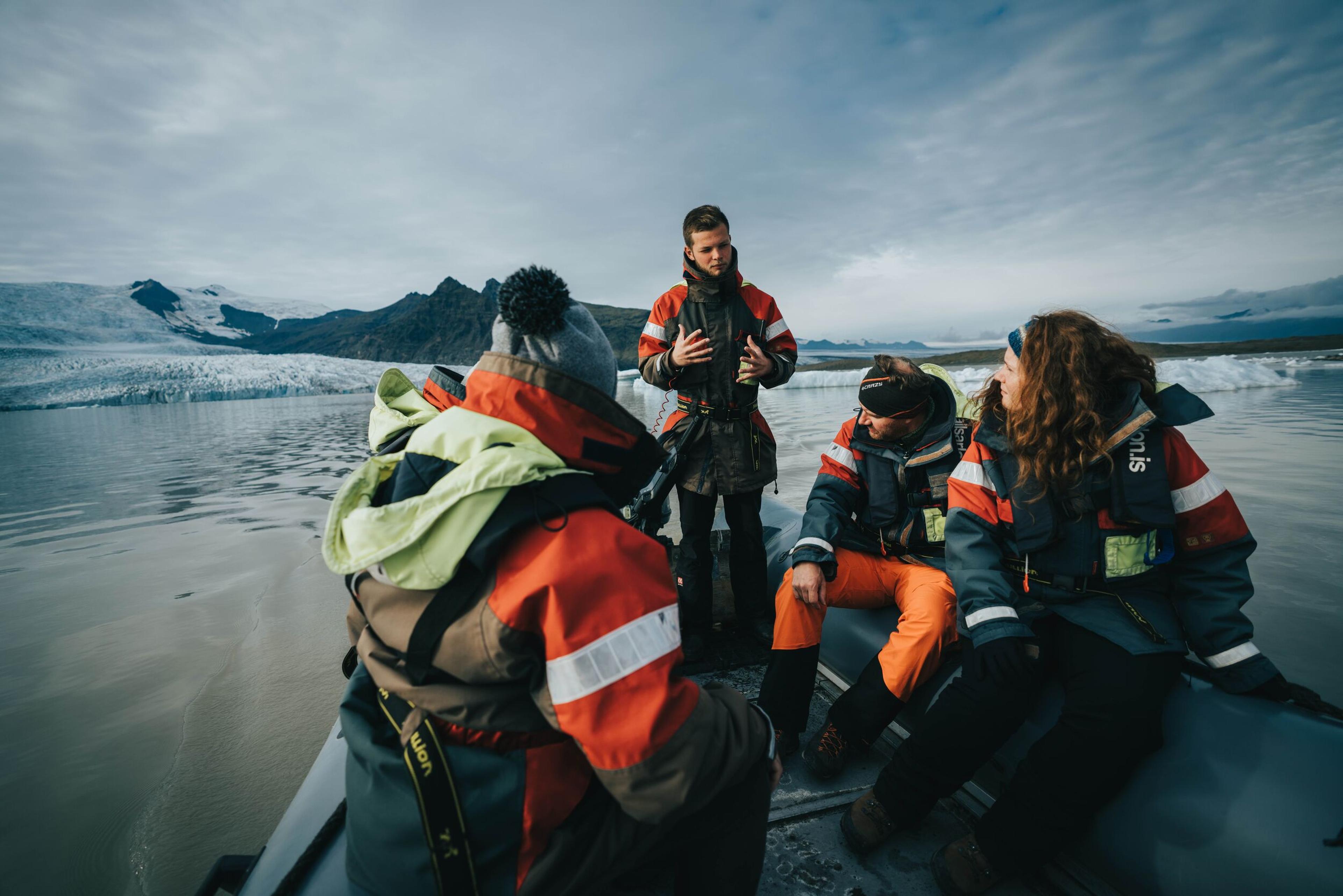 "A guide in a survival suit speaks to guests seated on an inflatable boat, with icebergs and mountains surrounding the glacial lagoon