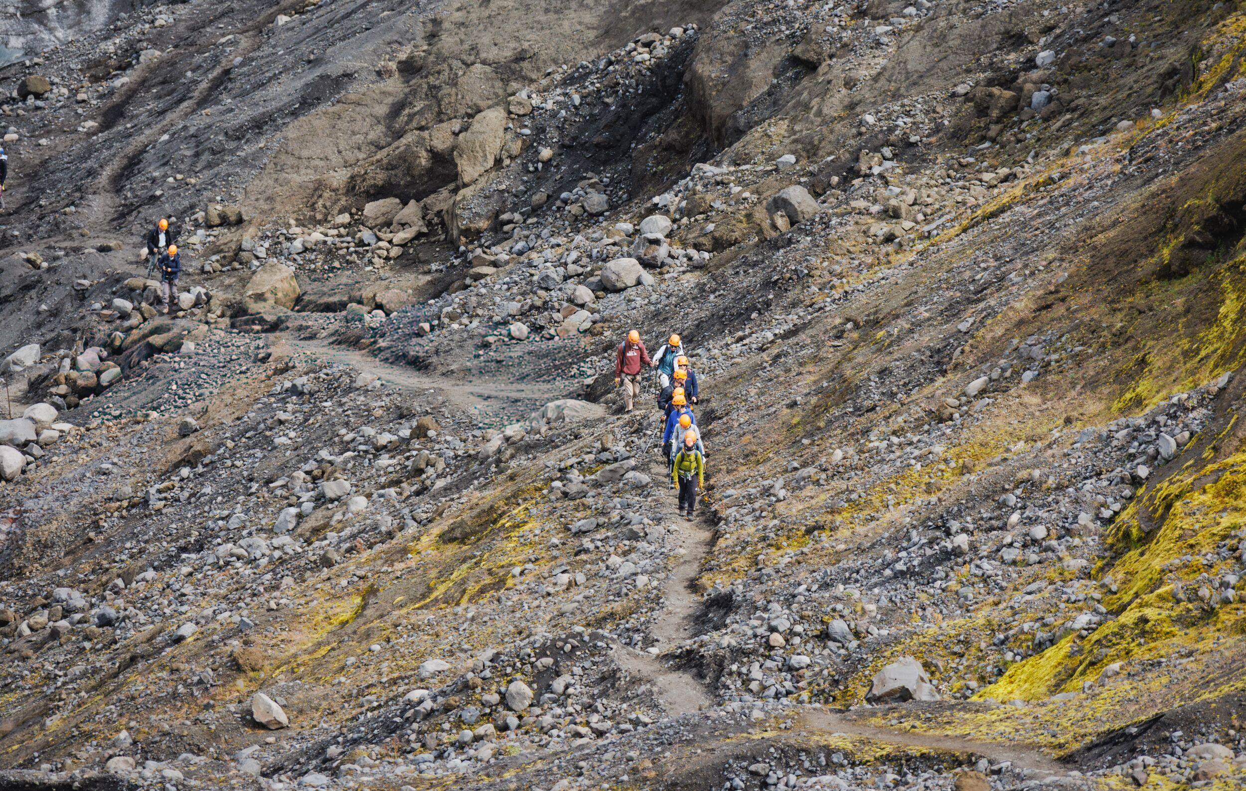 A line of hikers in orange helmets follows a winding trail down a rocky, sulfur-streaked volcanic slope.