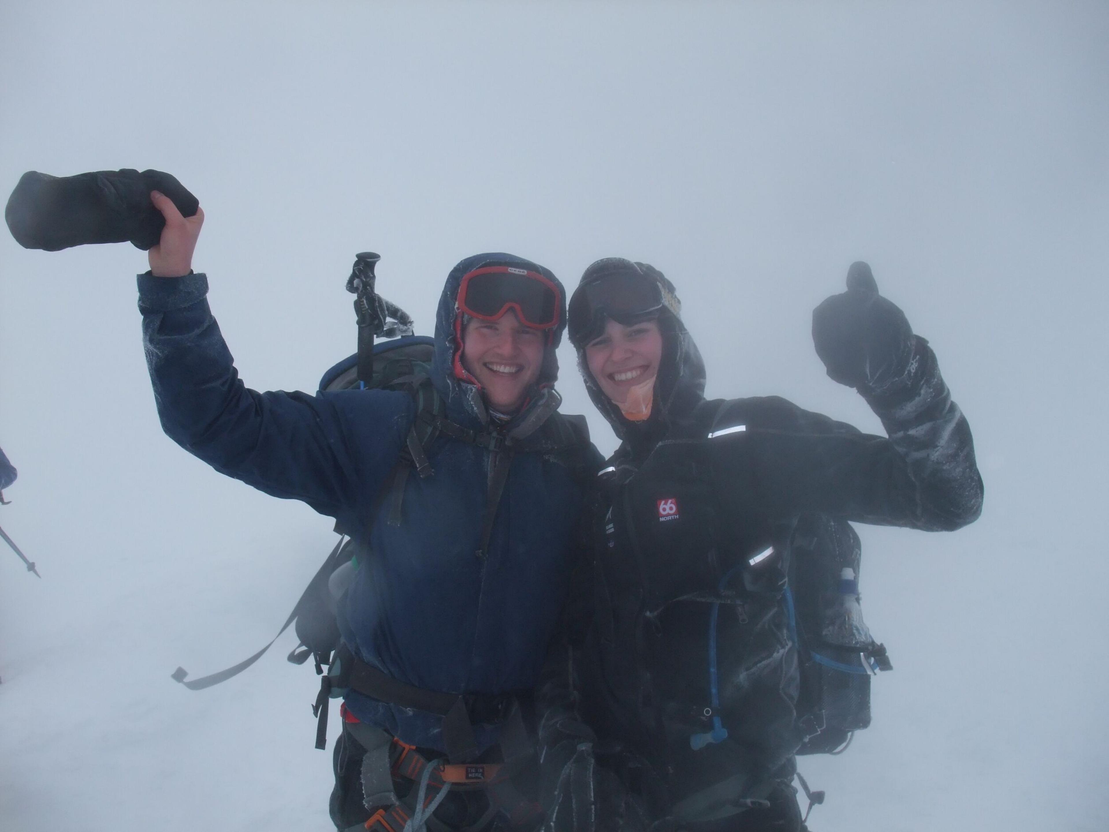 Two smiling hikers in snow-covered gear celebrate amidst foggy, challenging conditions on Eyjafjallajökull, one giving a thumbs-up and the other holding gear triumphantly.