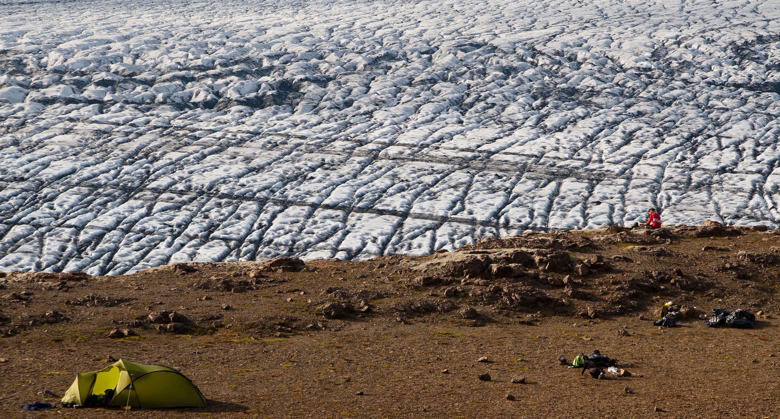 A green tent is pitched on rocky ground near the edge of a massive glacier in the Icelandic Highlands, highlighting the remote and rugged camping experience.